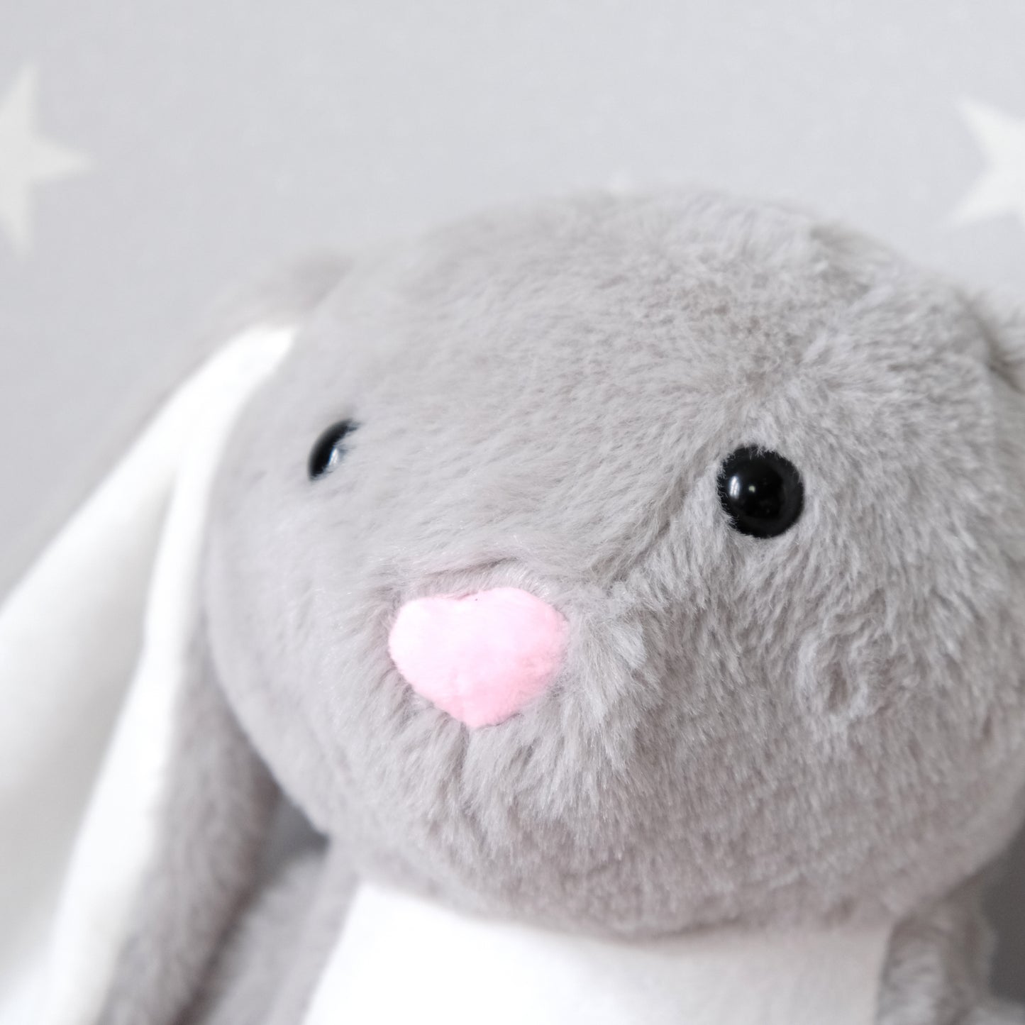 A close-up of a soft gray plush bunny toy with long white ears, black bead eyes, and a pink heart-shaped nose, set against a light gray background with faint white star shapes.