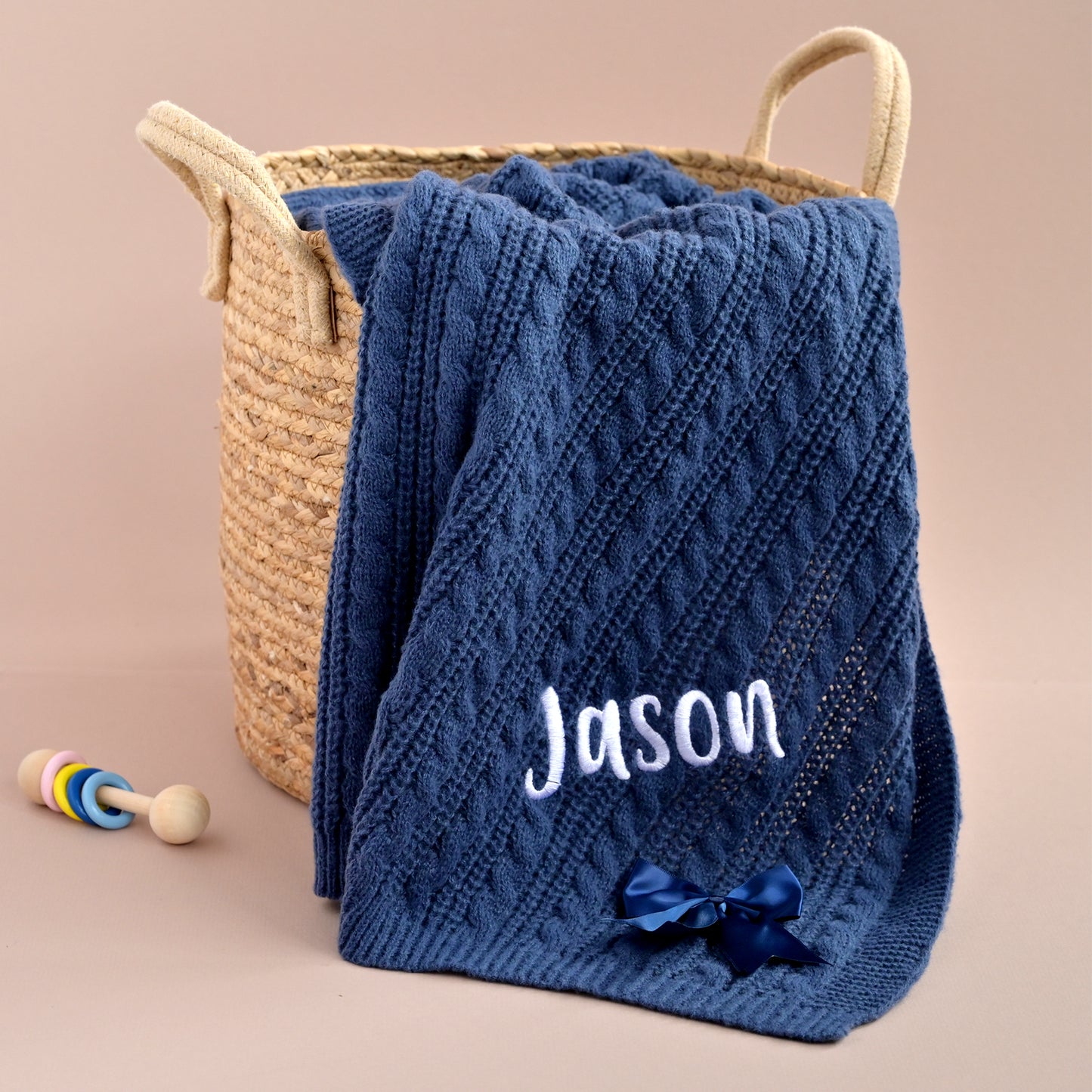 A navy blue cable-knit baby blanket with the name 'Jason' embroidered in white and a dark blue bow, draped over a woven straw basket on a beige background.