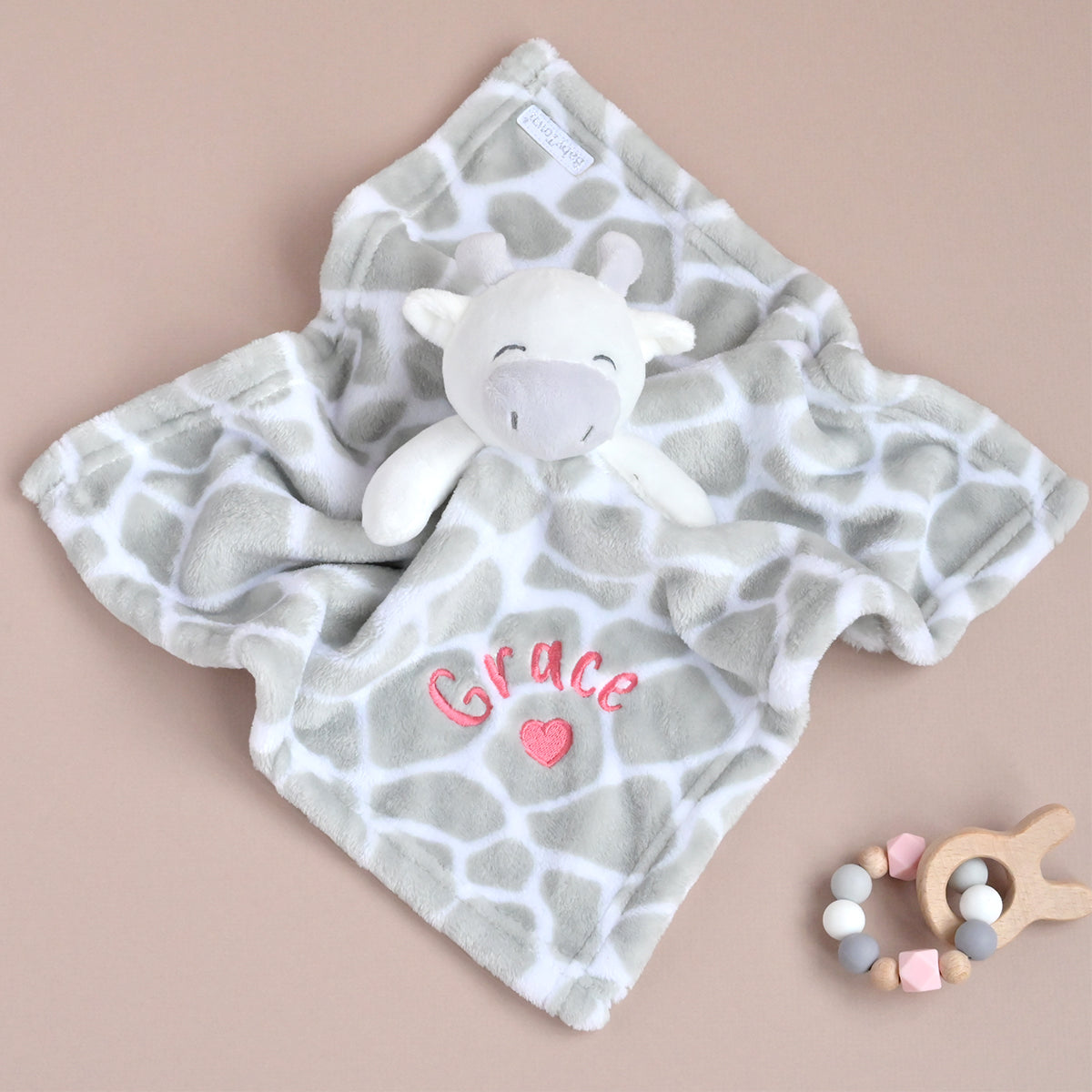 A personalized baby lovey blanket featuring a gray and white giraffe print, a plush white cow comforter, and pink embroidered text reading 'Grace' with a small pink heart. A wooden bead teether is placed on the right side, all arranged on a solid beige background.