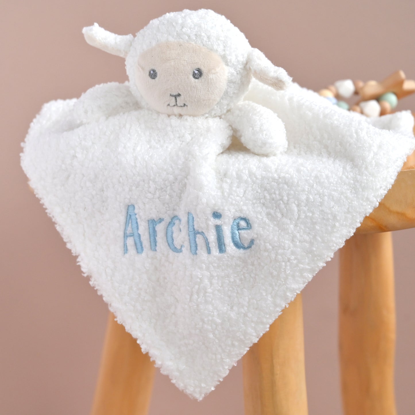 A white plush sheep lovey blanket with a soft teddy face, draped over a wooden stool. The front of the blanket is personalized with the name 'Archie' embroidered in light blue.