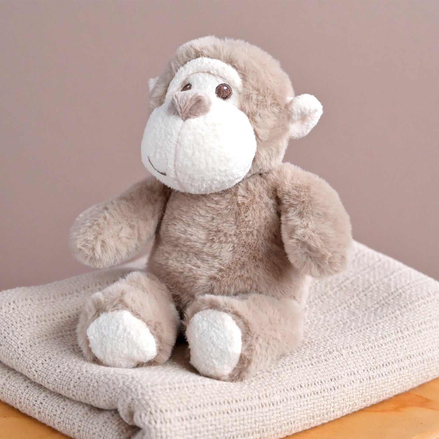 A plush stuffed monkey toy in light brown and white, sitting on a folded cream knit blanket against a soft beige background.