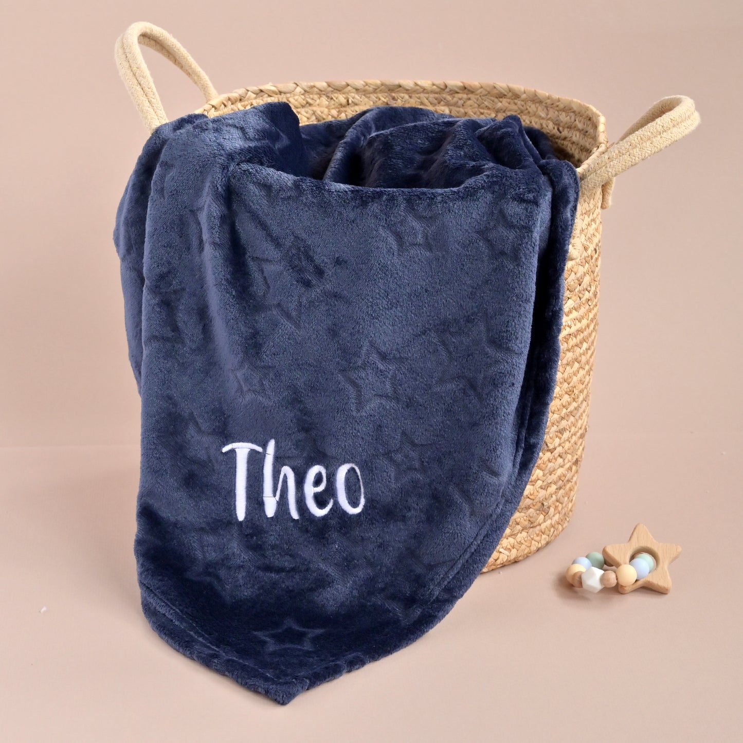 Navy blue blanket with 'Theo' embroidered on it, draped over a woven basket on a beige background.