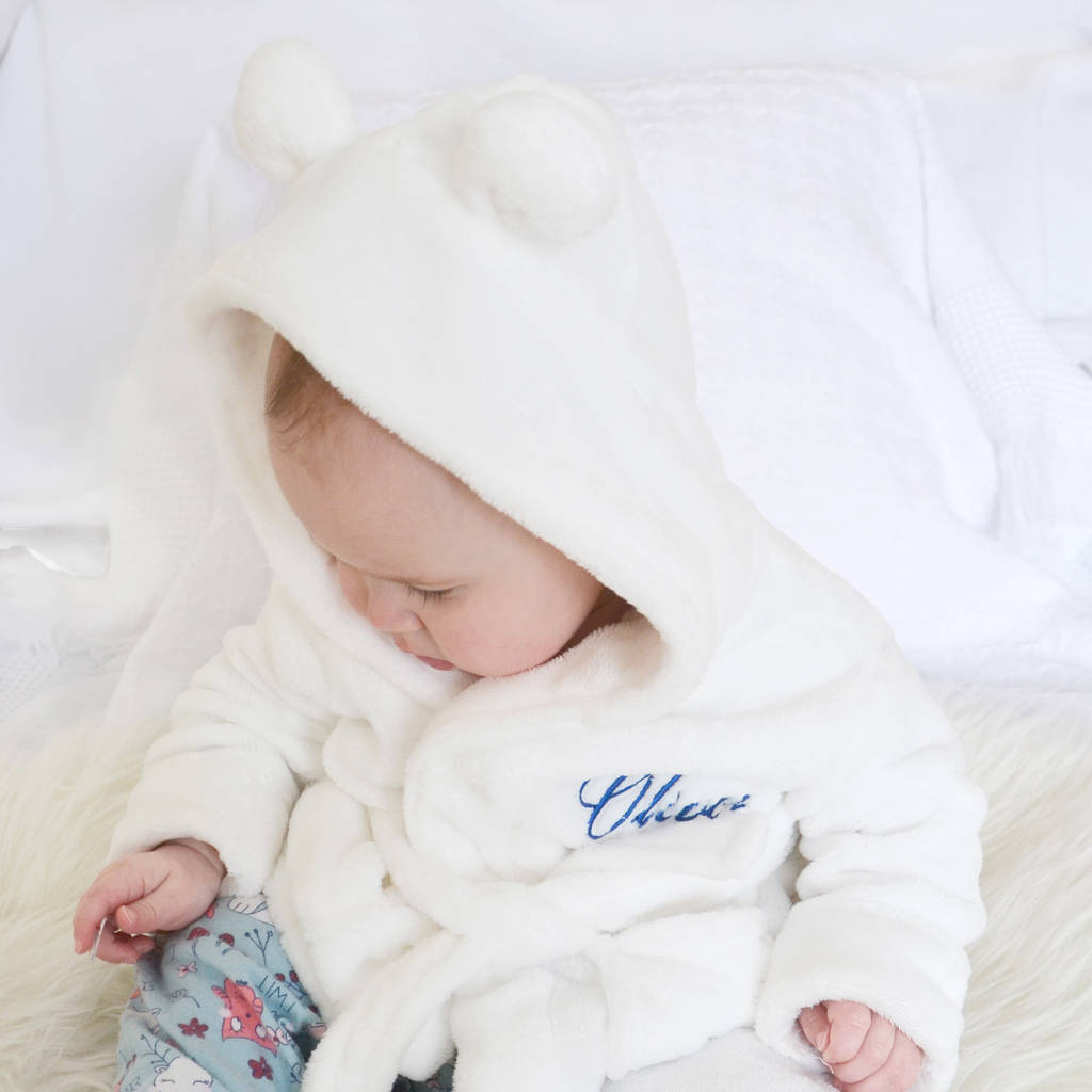 Baby wearing a white hooded dressing gown with bear ears and a small embroidered elephant, sitting on a woven rug in a bright nursery.