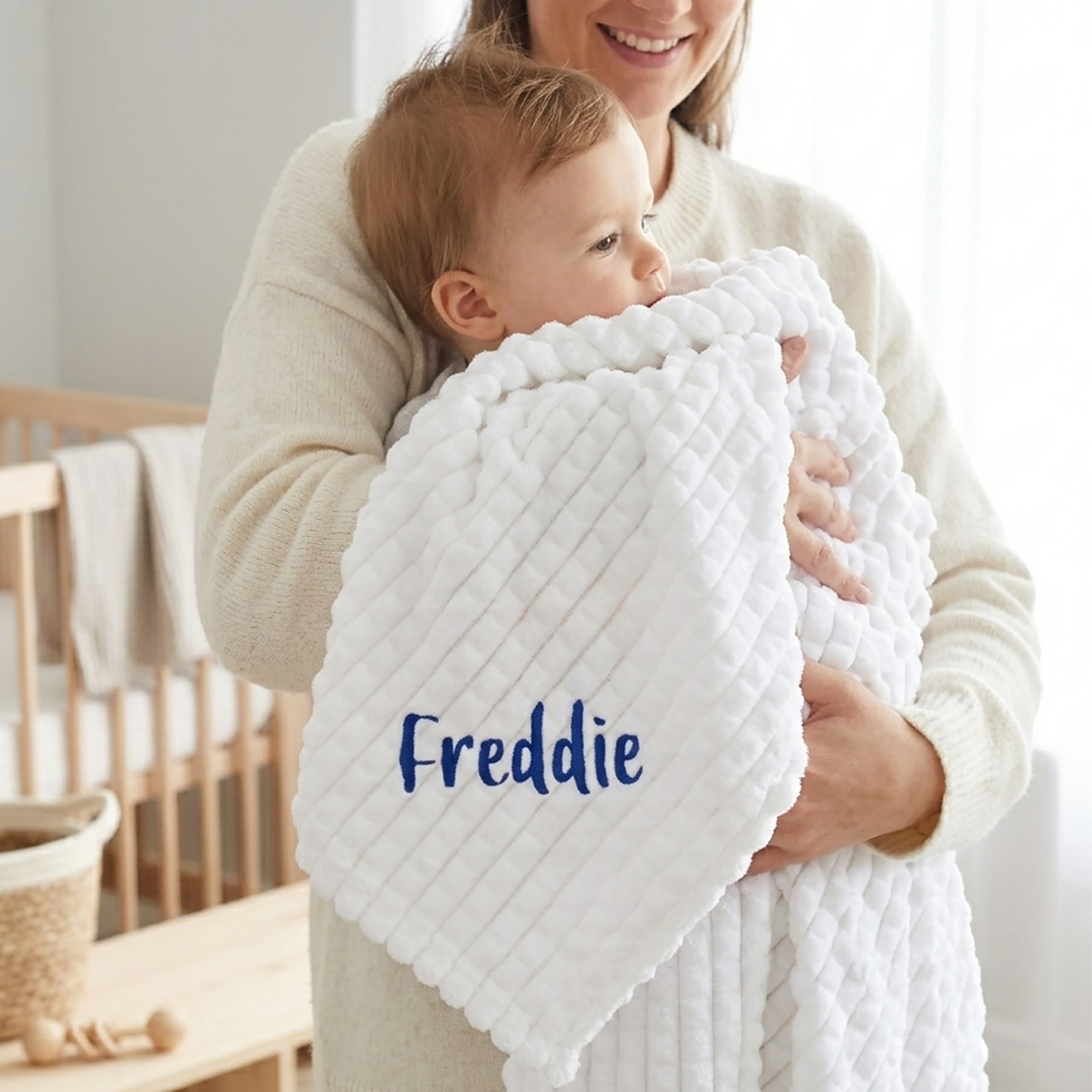 White ribbed fleece baby blanket personalised with the name Freddie in navy embroidery, held by a mother cradling her baby.