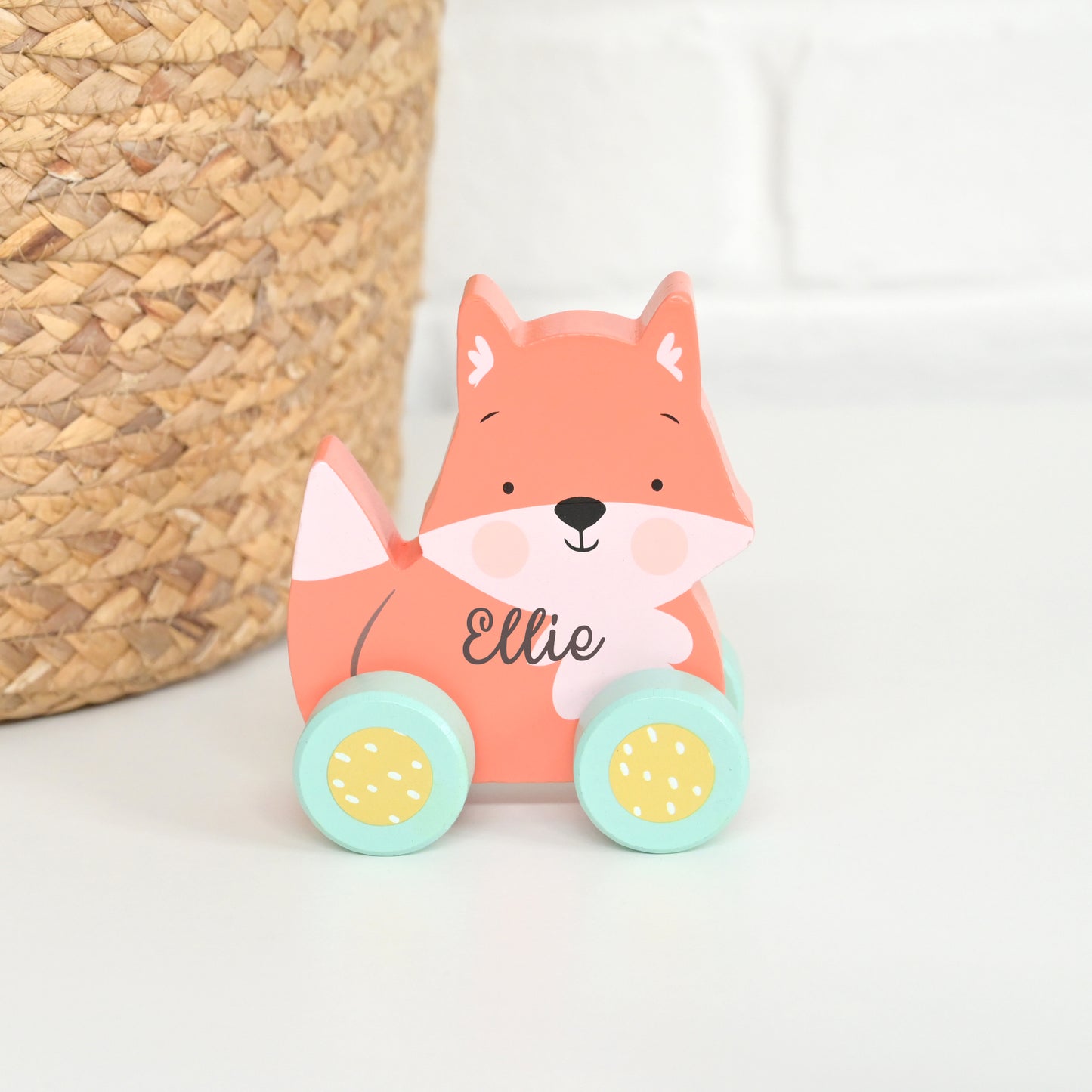 A personalized wooden toy car shaped like a fox, with the name 'Ellie' written on its chest. The toy features a salmon-pink fox body, a white muzzle and chest, mint green wheels with yellow centers, and is styled next to a woven basket on a white surface.