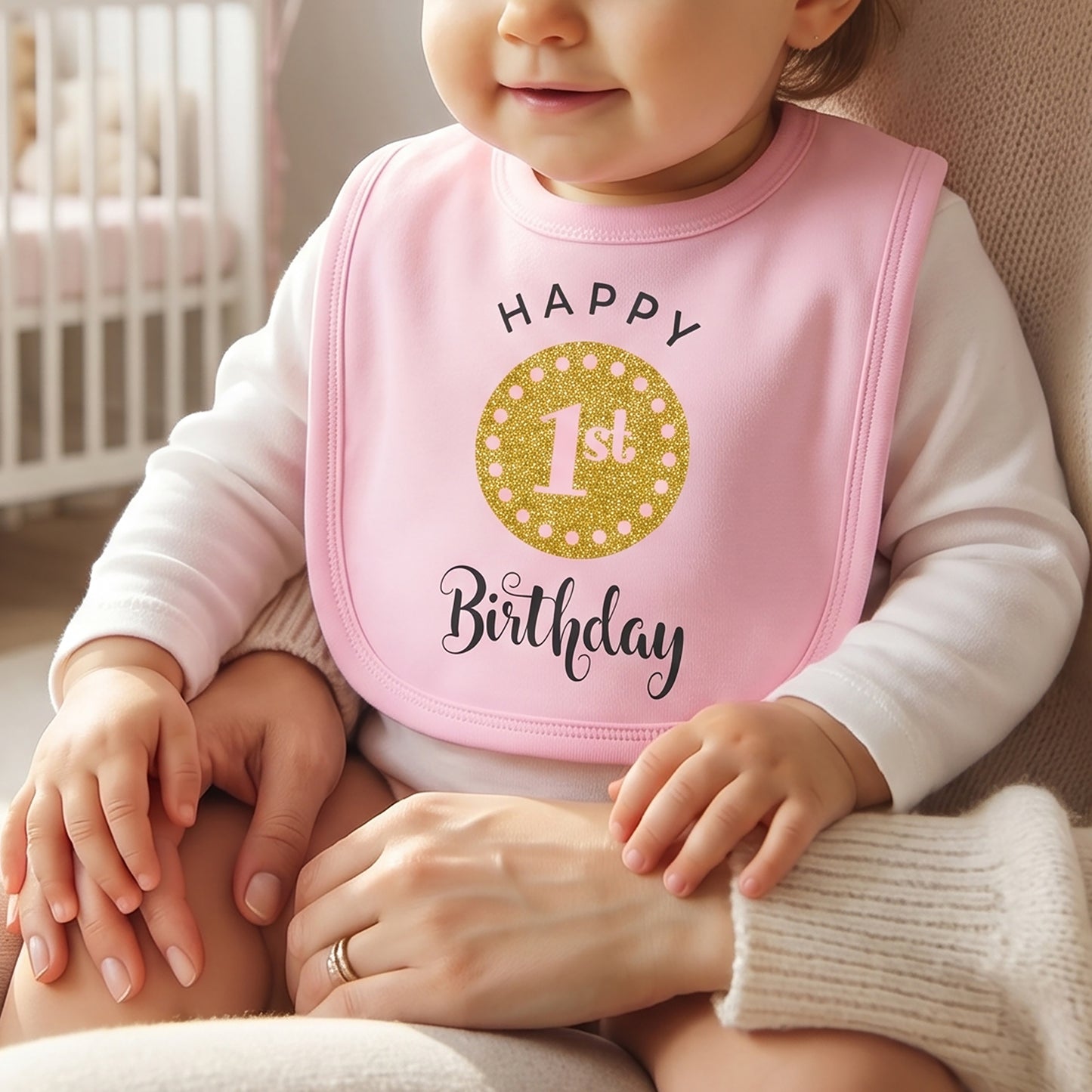 Pink baby bib printed with "Happy 1st Birthday" in black and gold glitter lettering, worn by a baby sitting on a caregiver's lap.