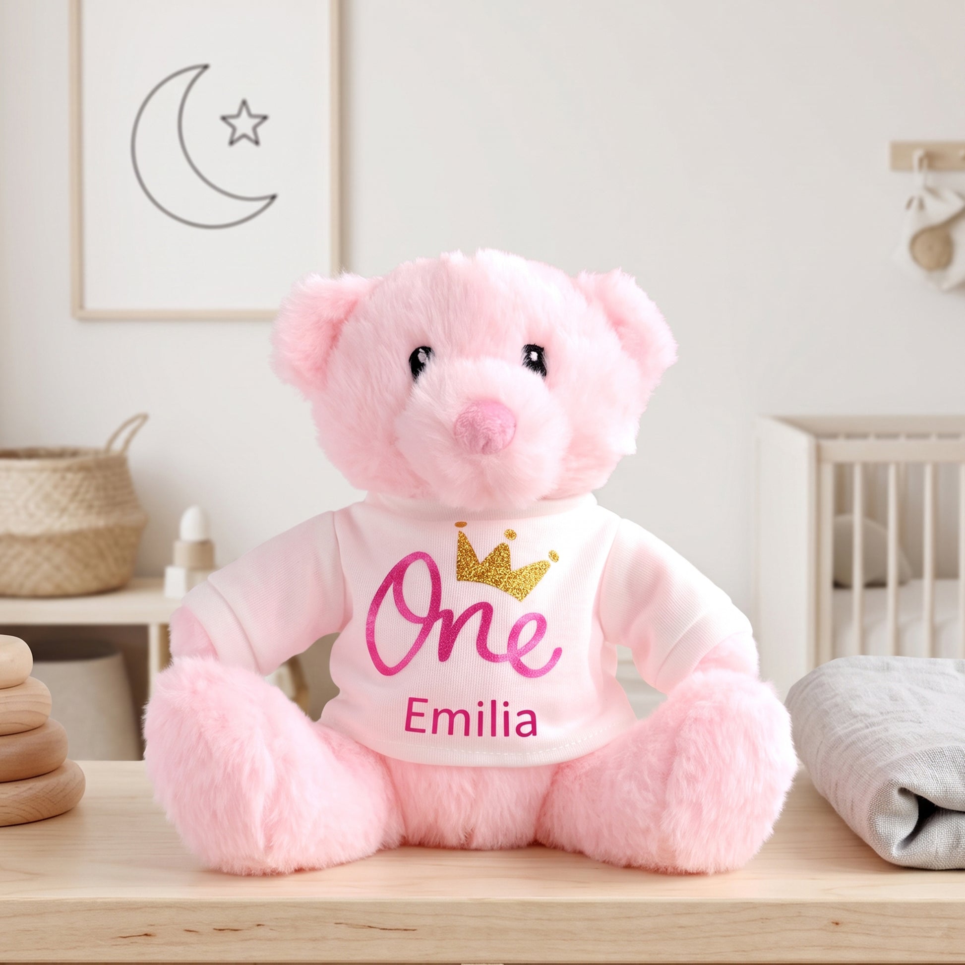 A pink plush teddy bear sitting on a wooden surface in a softly styled nursery-like room. The bear wears a white shirt with the word "One" in pink script, a small gold crown graphic, and the name "Emilia" in pink text.
