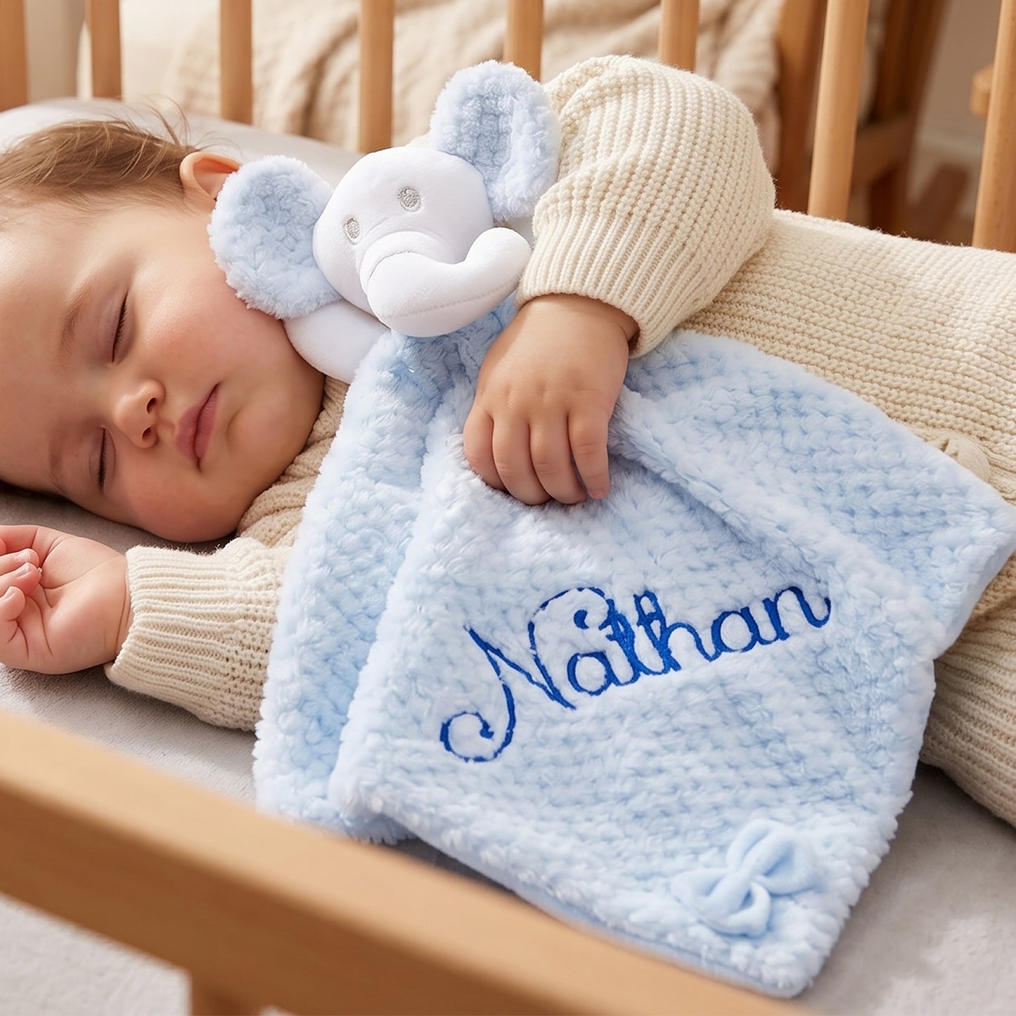 A sleeping baby tucked in a wooden crib, wrapped in a light blue waffle-knit security blanket with an elephant toy and the name 'Nathan' embroidered in blue.