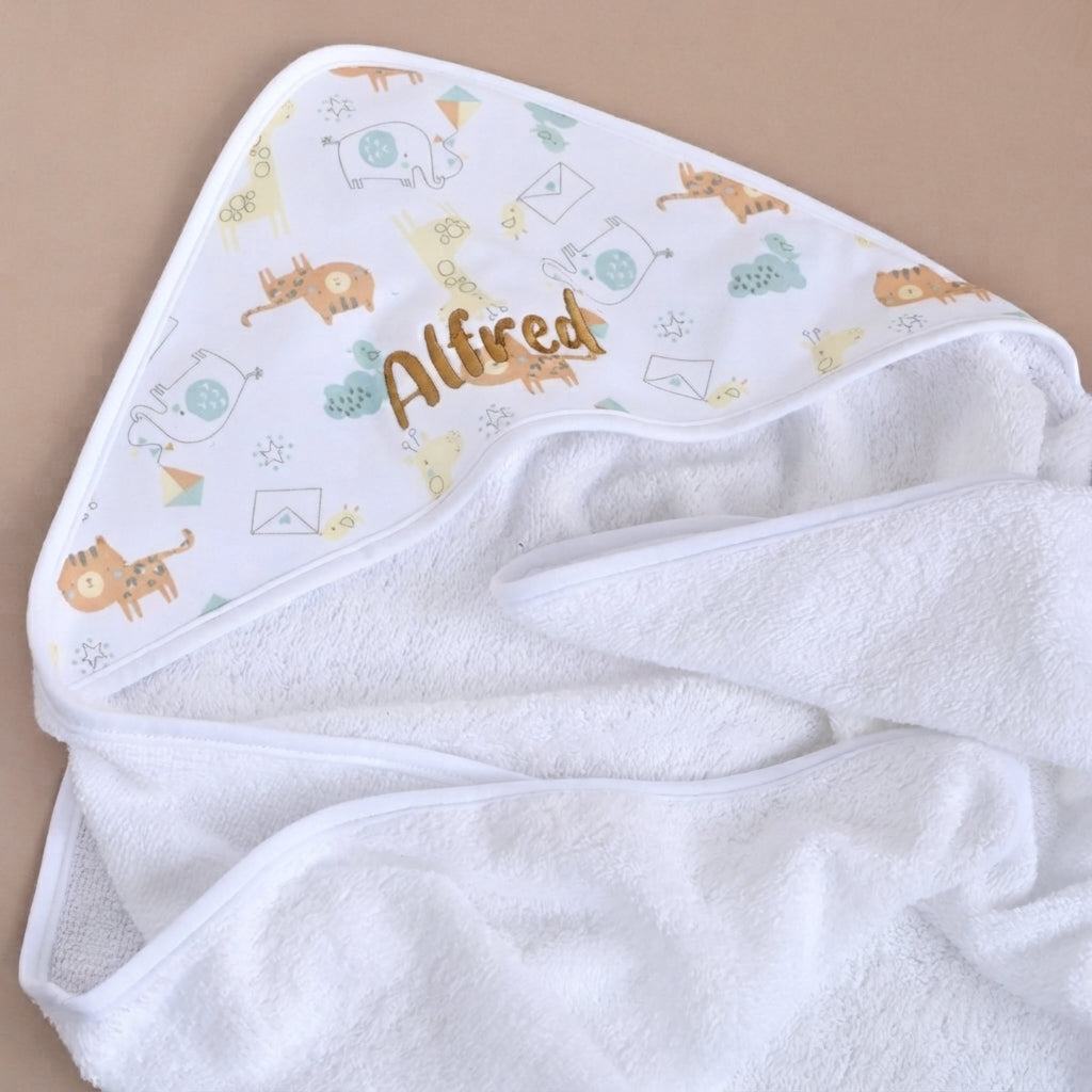 White hooded baby towel with a safari animal print in gold and blue, personalised with the name Alfred embroidered in gold on the hood, worn by a smiling baby.