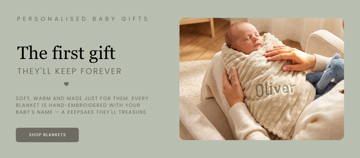 Baby wrapped in a blanket with embroidered name, held by an adult, on a light background.