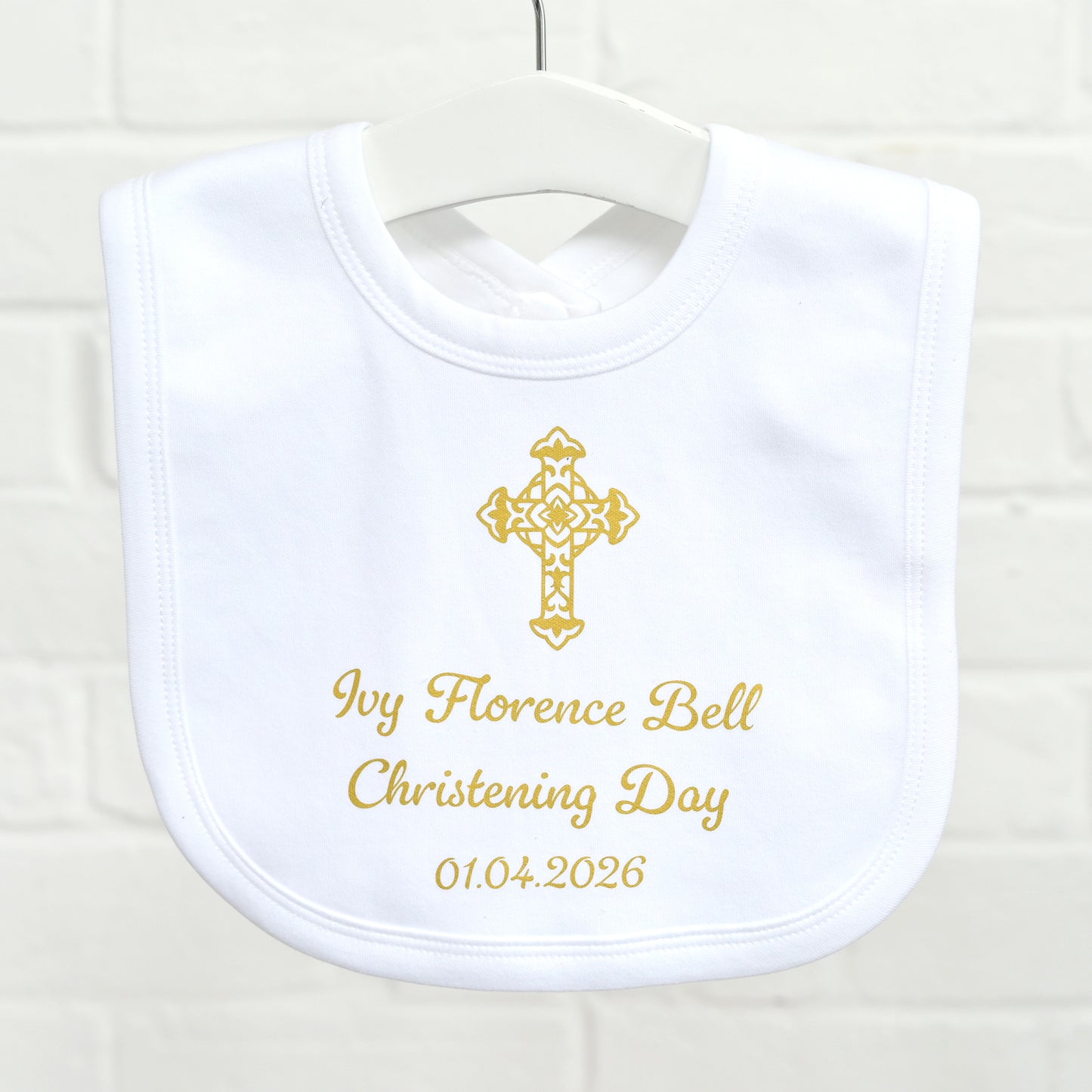 A white baby bib with gold embroidery featuring a cross and the personalized text 'Ivy Florence Bell Christening Day 01.04.