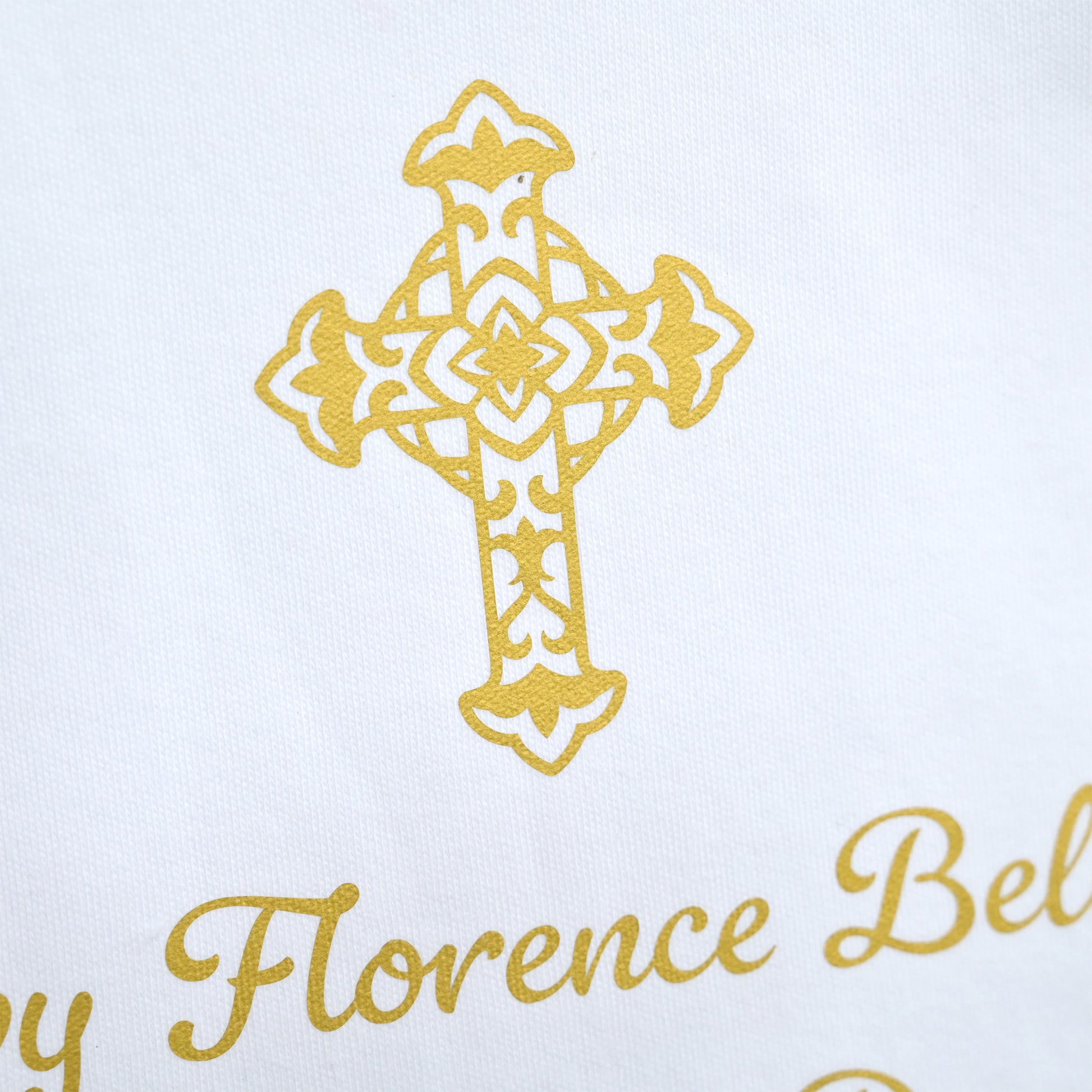 Close-up of a white textile featuring a gold embroidered cross and partial gold script text reading 'by Florence Bel'.