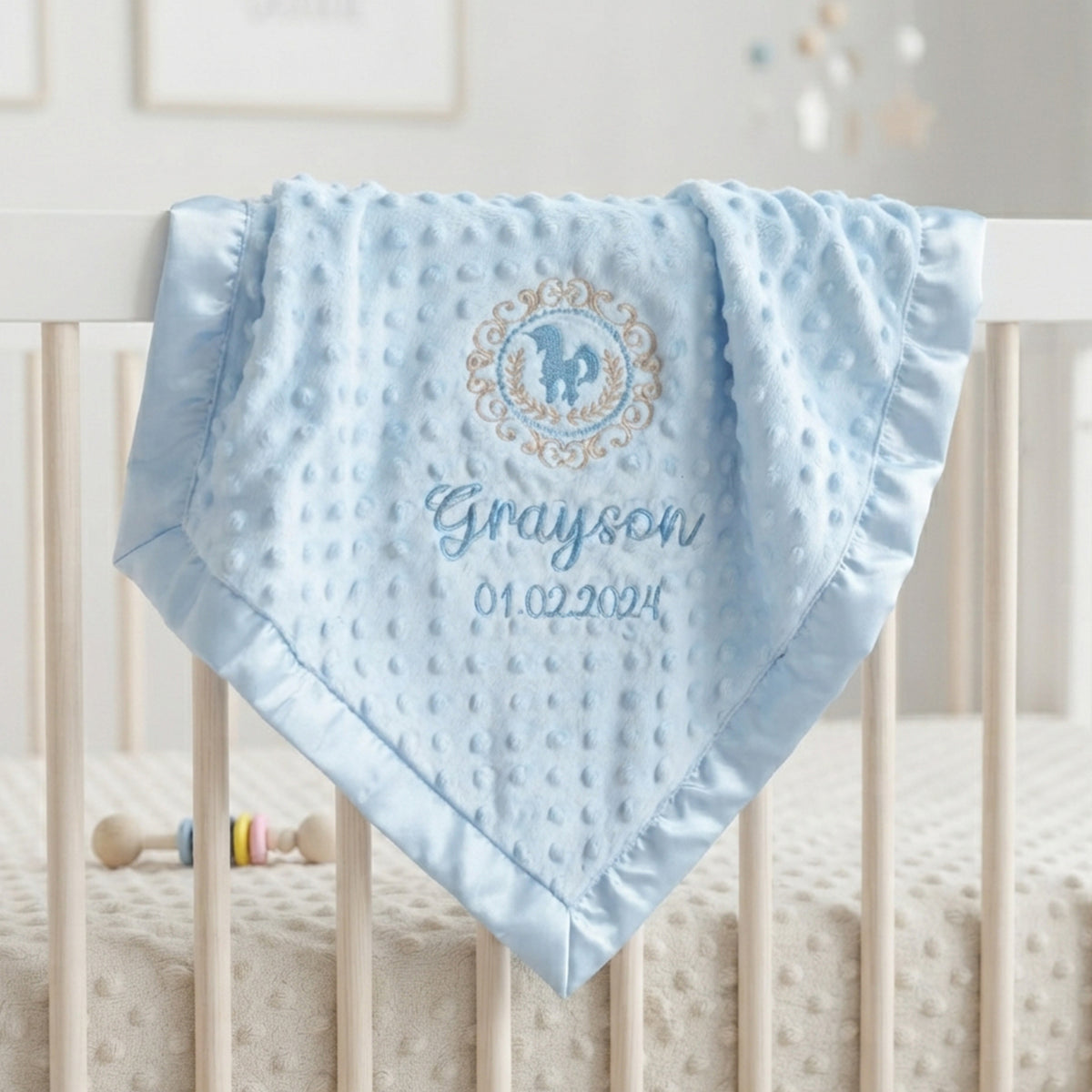 A light blue minky dot baby blanket with a personalized monogram and name 'Grayson' draped over the railing of a white crib.