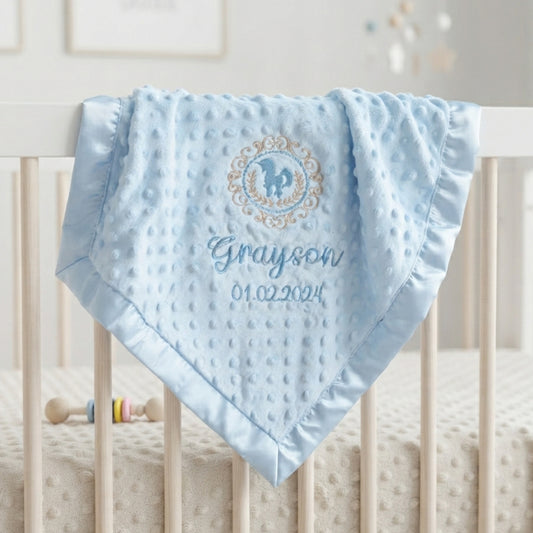 A light blue minky dot baby blanket with a personalized monogram and name 'Grayson' draped over the railing of a white crib.