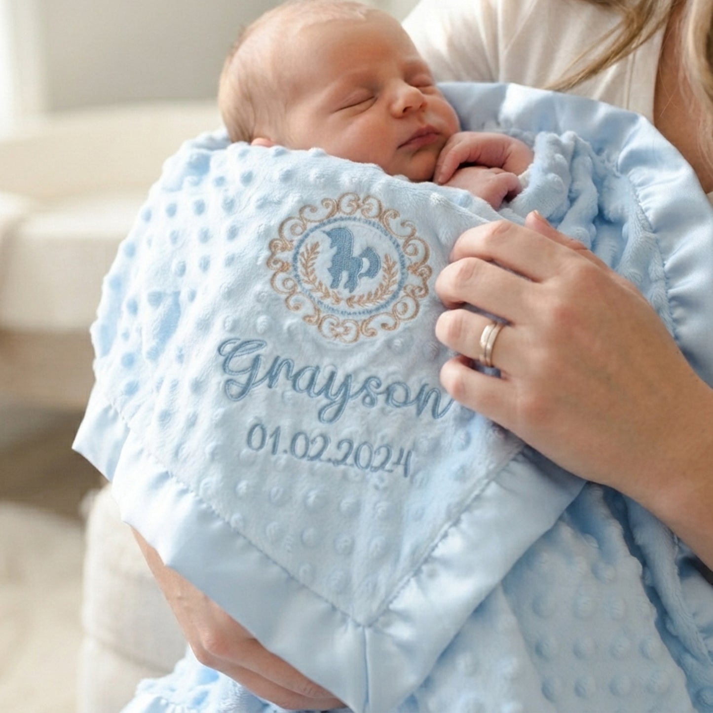 A sleeping newborn baby wrapped in a light blue textured muslin swaddle blanket held by a mother. The blanket features a personalized circular monogram with a horse and decorative border, and the name 'Grayson' with the date '01.