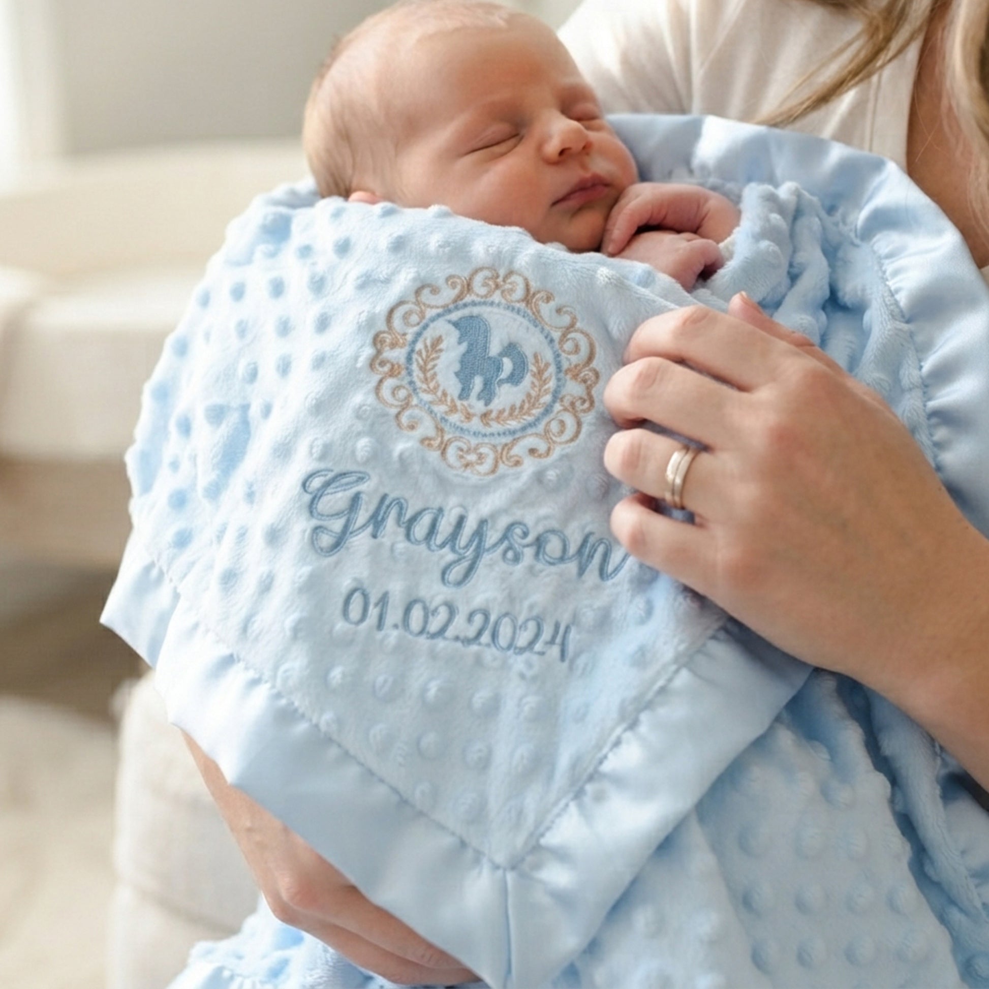 A sleeping newborn baby wrapped in a light blue textured muslin swaddle blanket held by a mother. The blanket features a personalized circular monogram with a horse and decorative border, and the name 'Grayson' with the date '01.