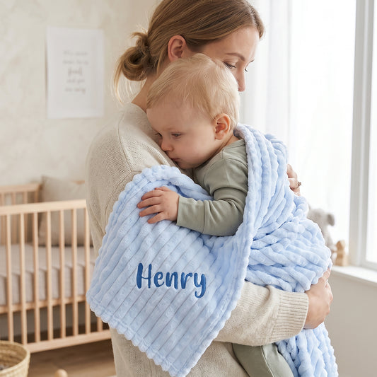 Blue ribbed fleece baby blanket personalised with the name Henry in navy embroidery, wrapped around a baby being held by his mother.