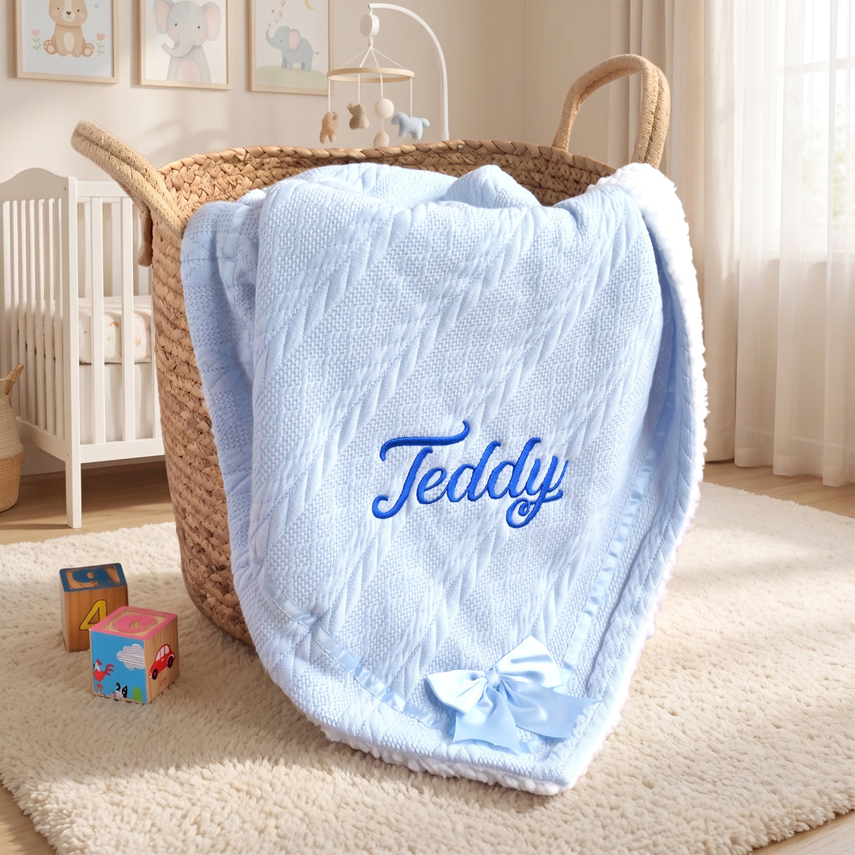 A light blue textured blanket with the name 'Teddy' embroidered in blue script and a matching blue bow, draped over a woven basket in a softly lit nursery.