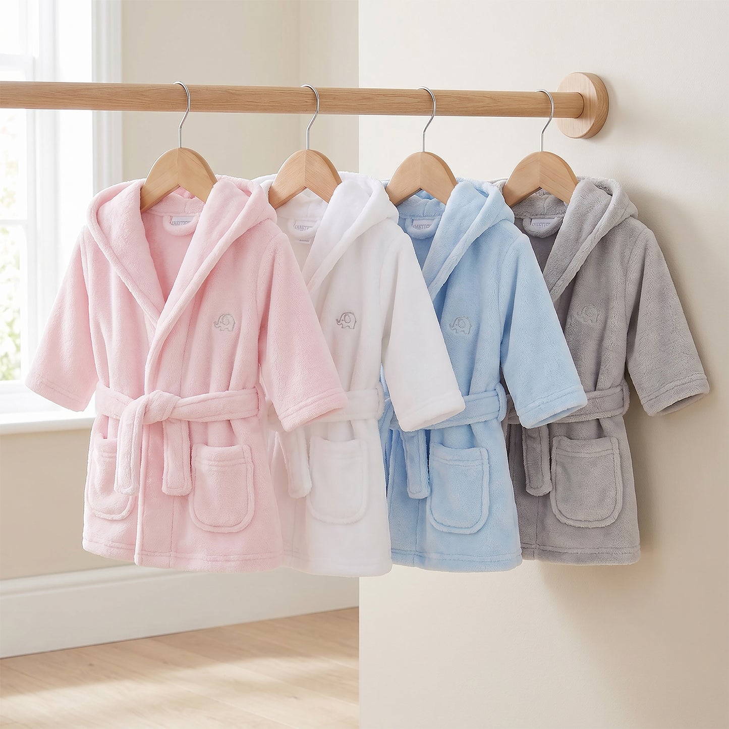 Four children's hooded bathrobes in pink, white, light blue, and gray hang on wooden hangers from a wooden clothing rack against a beige wall.