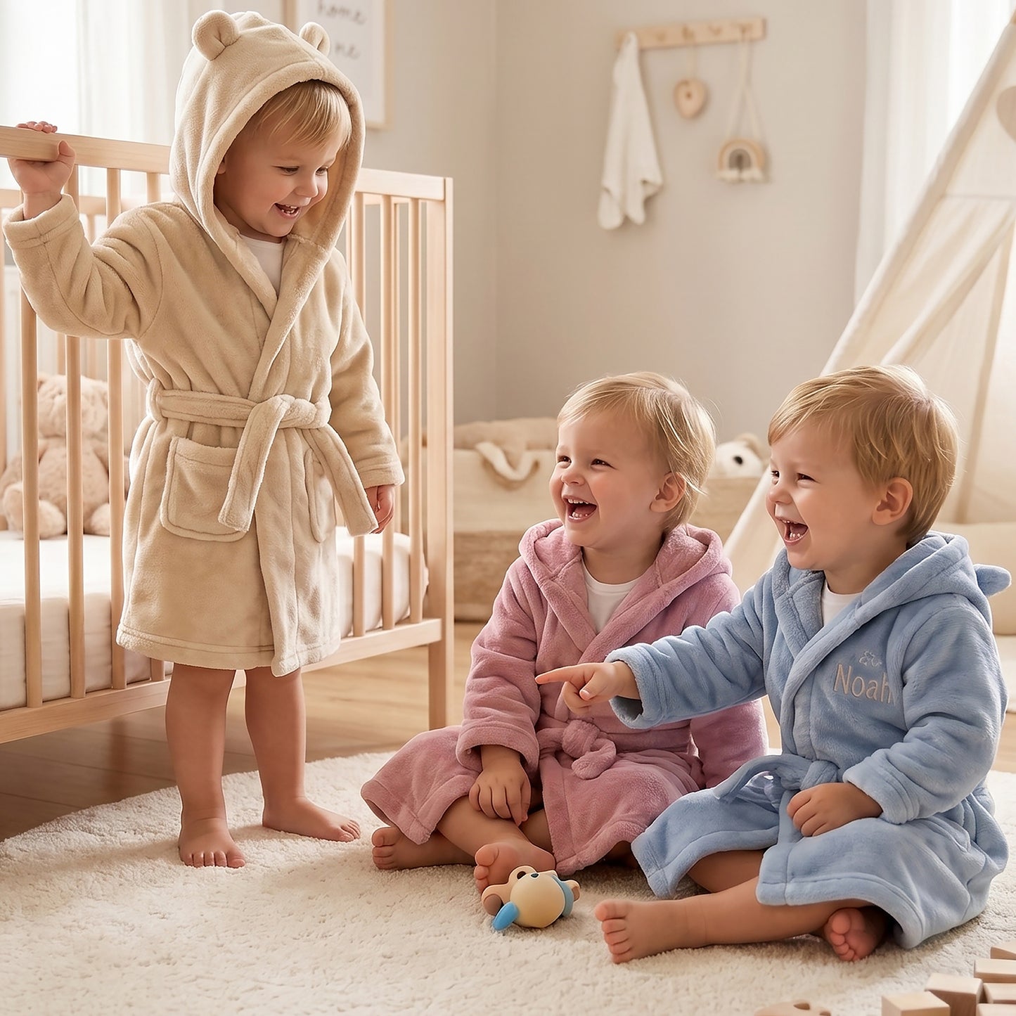 Three toddlers wearing plush hooded bathrobes in a cozy nursery setting. One child stands by a wooden crib in a beige robe, while two other children sit on a white rug on the floor in pink and light blue robes.