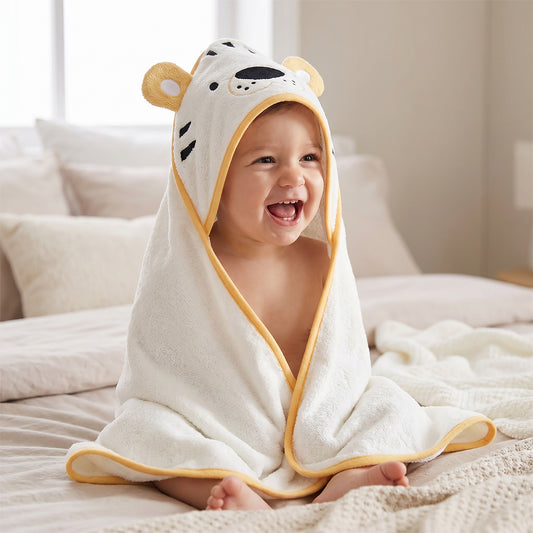 Cream hooded baby towel shaped like a tiger cub face with mustard yellow ears, worn by a laughing baby sitting on a bed.