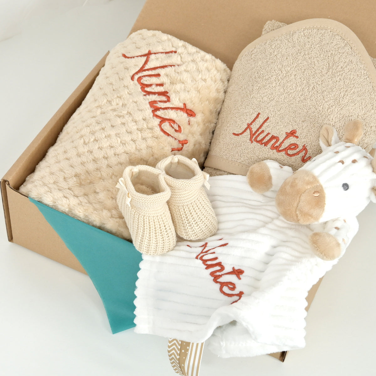 A baby gift set in a brown cardboard box, featuring a cream waffle-knit blanket and a matching bath towel embroidered with 'Hunter' in red, a pair of cream knit booties, a white striped muslin baby wrap with red embroidery, and a plush cow toy.