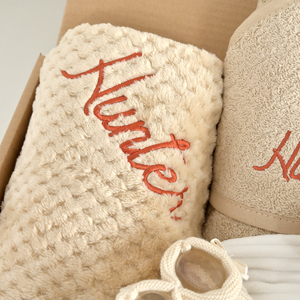 A close-up of a gift set containing a cream-colored textured blanket with red embroidered script reading 'Hunter' and a matching beige towel, both placed in a brown cardboard box.