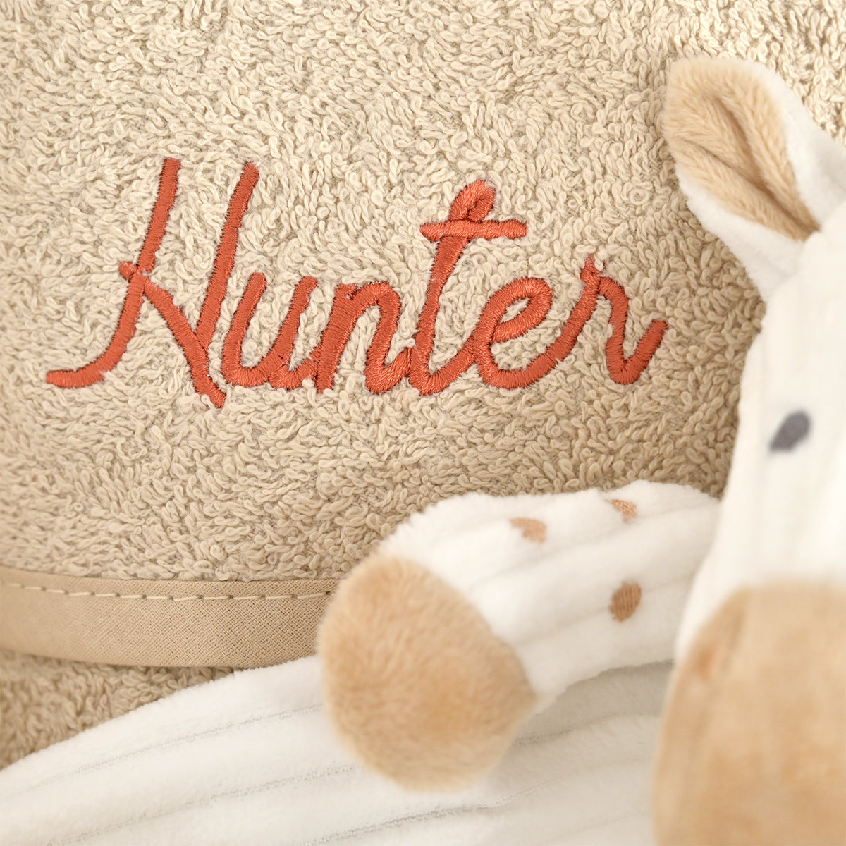 Close-up of a beige textured blanket or towel with the name 'Hunter' embroidered in orange script, alongside a white plush toy with tan spots.