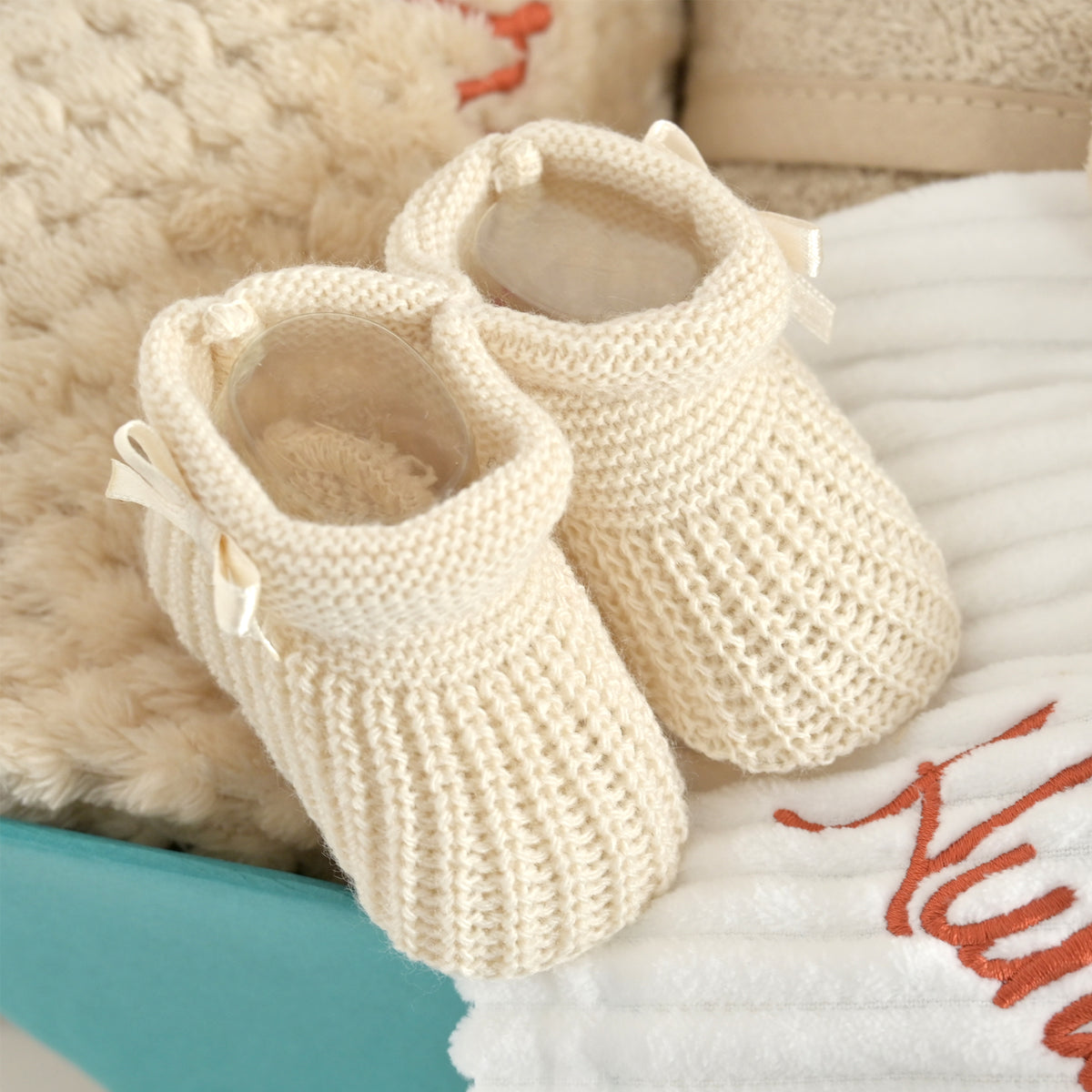A pair of cream-colored, ribbed-knit baby booties with small bow accents, styled on a white blanket with red embroidery next to a plush beige toy.