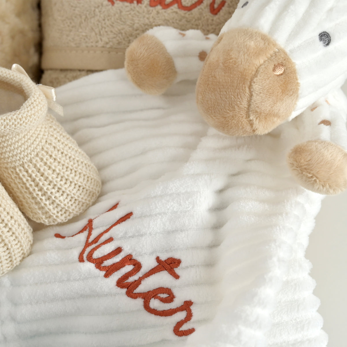 A close-up of a white ribbed plush blanket with the name 'Hunter' embroidered in brown script, styled with a beige knit bootie and a plush sheep toy.