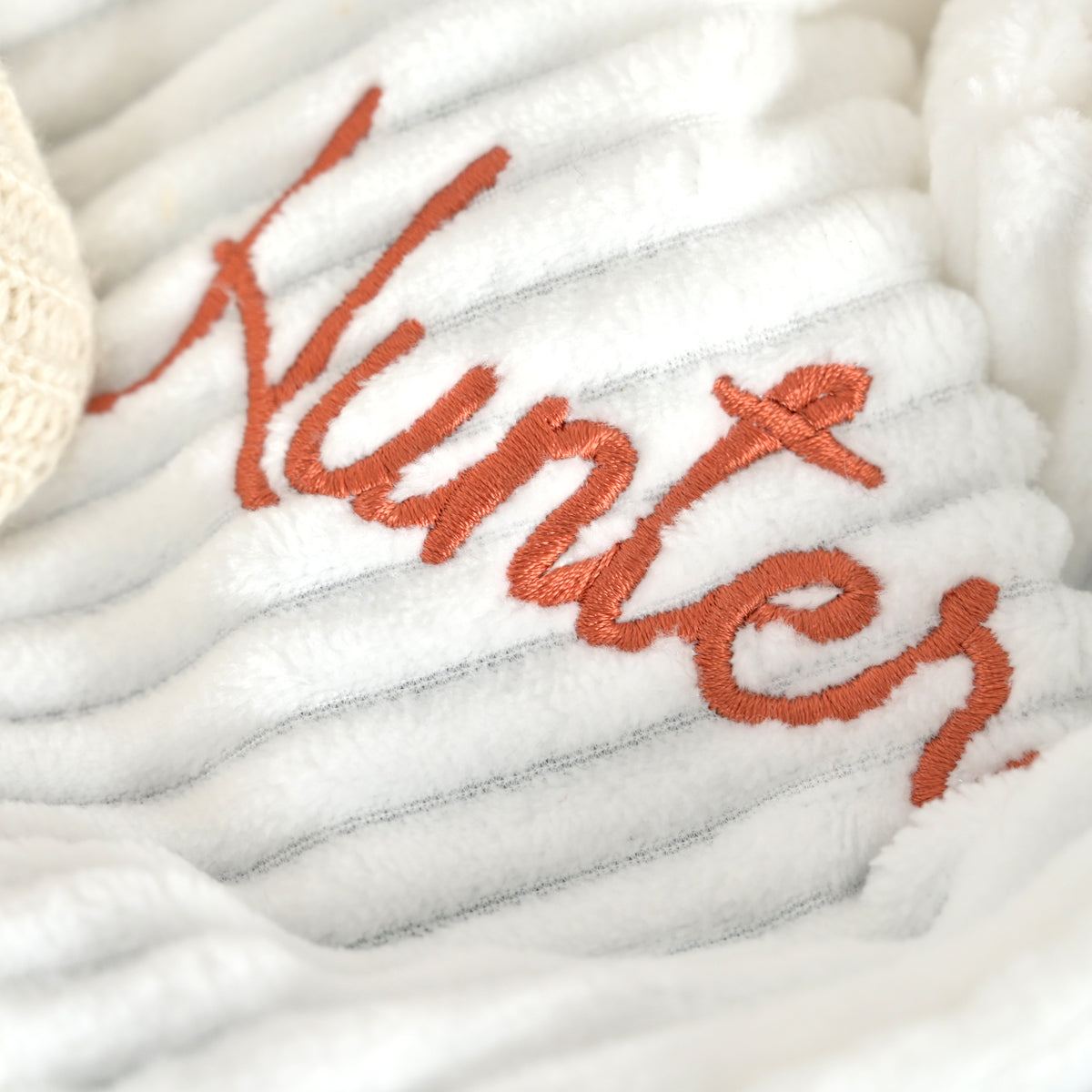 Close-up of a white plush ribbed blanket with orange embroidered script reading 'Hunter' and a small white knit item partially visible at the left edge.
