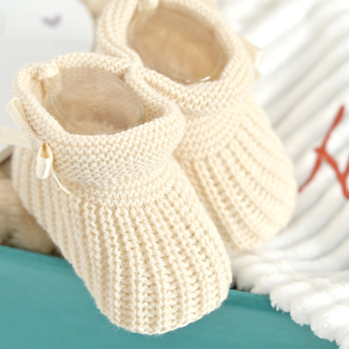 A pair of cream-colored, ribbed-knit baby booties with small fabric bows, displayed on a teal surface next to a white textured blanket.