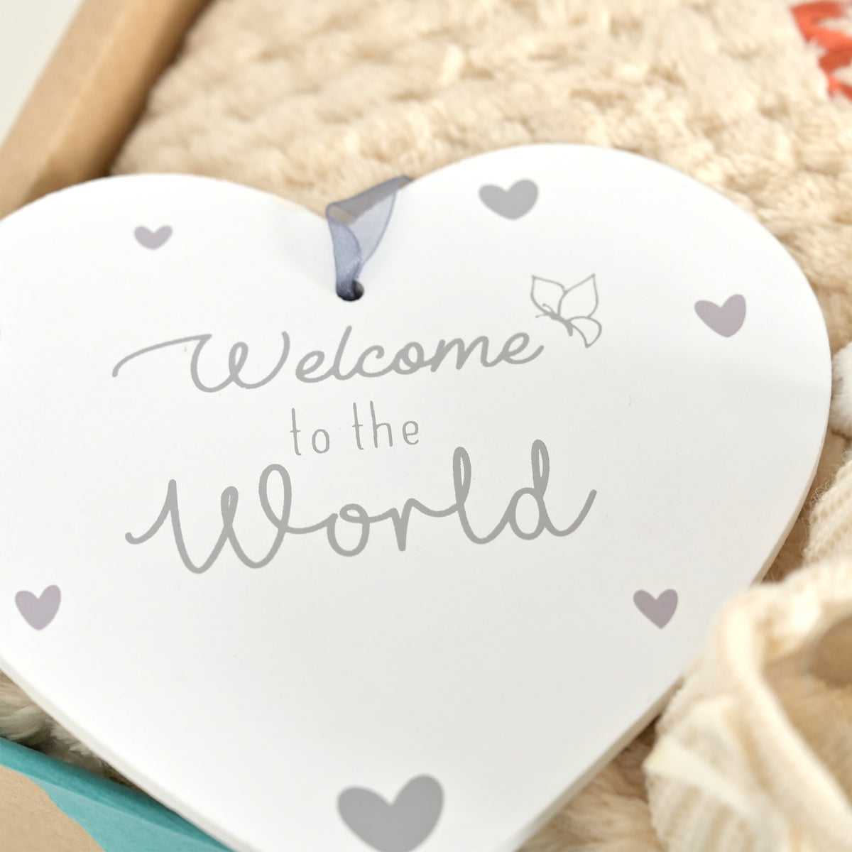 A white heart-shaped hanging ornament with the words 'Welcome to the World' in gray script and small gray heart accents, displayed on a soft cream textured blanket.