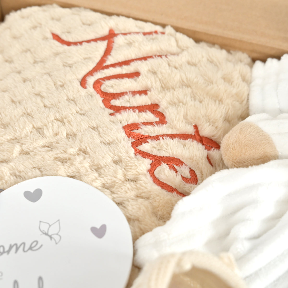 A close-up of a beige textured blanket with orange embroidered script reading 'Nurse', placed in a gift box alongside a white knit baby hat and a round white card with butterfly and heart details.