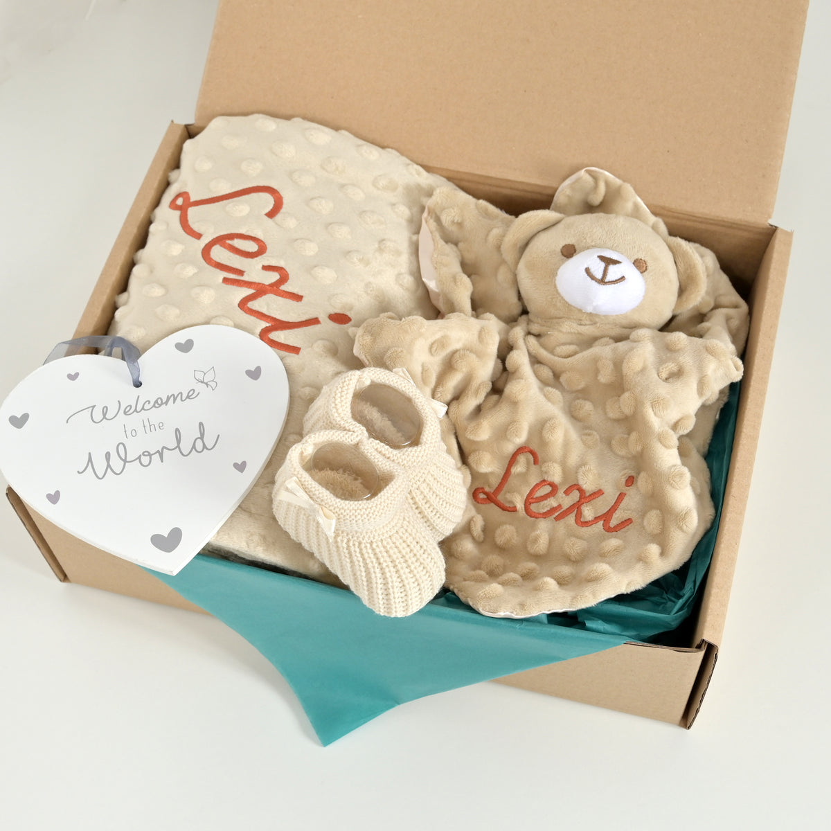 A personalized baby gift set in a brown cardboard box, featuring a plush teddy bear comforter, a textured beige blanket with 'Lexi' embroidered in red, a pair of cream knit booties, and a white heart-shaped tag that reads 'Welcome to the World'.