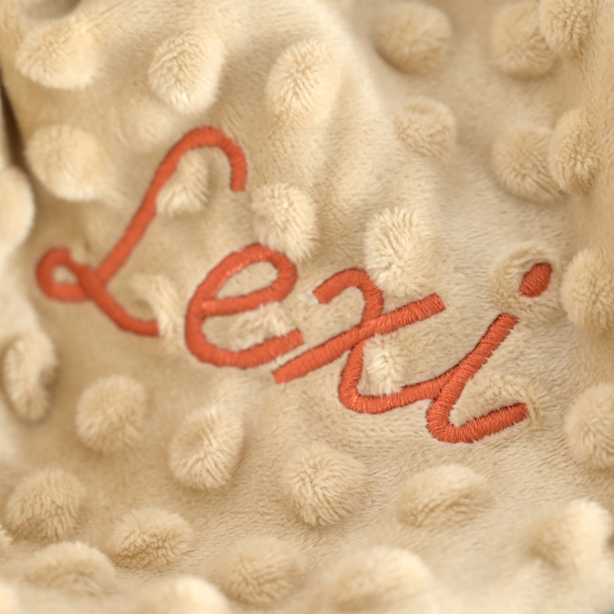 Close-up of a beige textured plush blanket with raised bumpy dots and the name 'Lexi' embroidered in orange-red script across the center.