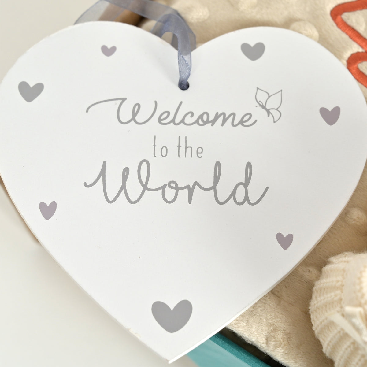 A white heart-shaped hanging sign with the words 'Welcome to the World' in gray script and small gray heart accents, shown hanging with a gray ribbon in a styled setting.