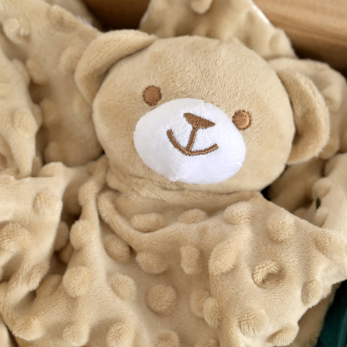 A close-up of a plush teddy bear comforter or lovey blanket with a soft tan body, white snout, embroidered brown face details, and a textured dotted blanket attached.