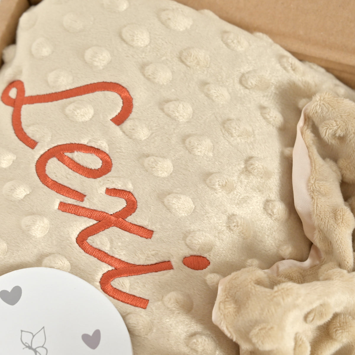 A close-up of a beige textured blanket with a raised dot pattern, featuring the word 'love' embroidered in orange script. A white circular tag with grey hearts is partially visible in the bottom-left corner, and the blanket is nestled inside a brown cardboard box.