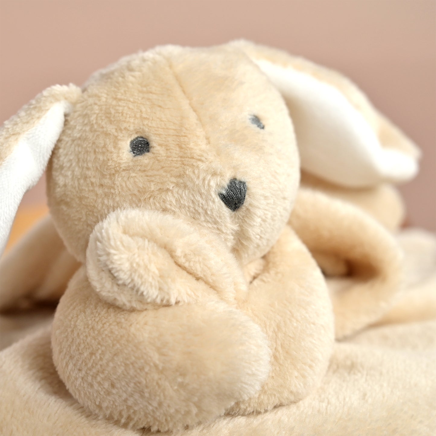 A close-up of a soft plush bunny toy or comforter in warm beige and cream tones, with embroidered black eyes and nose, resting against a pale pink background.