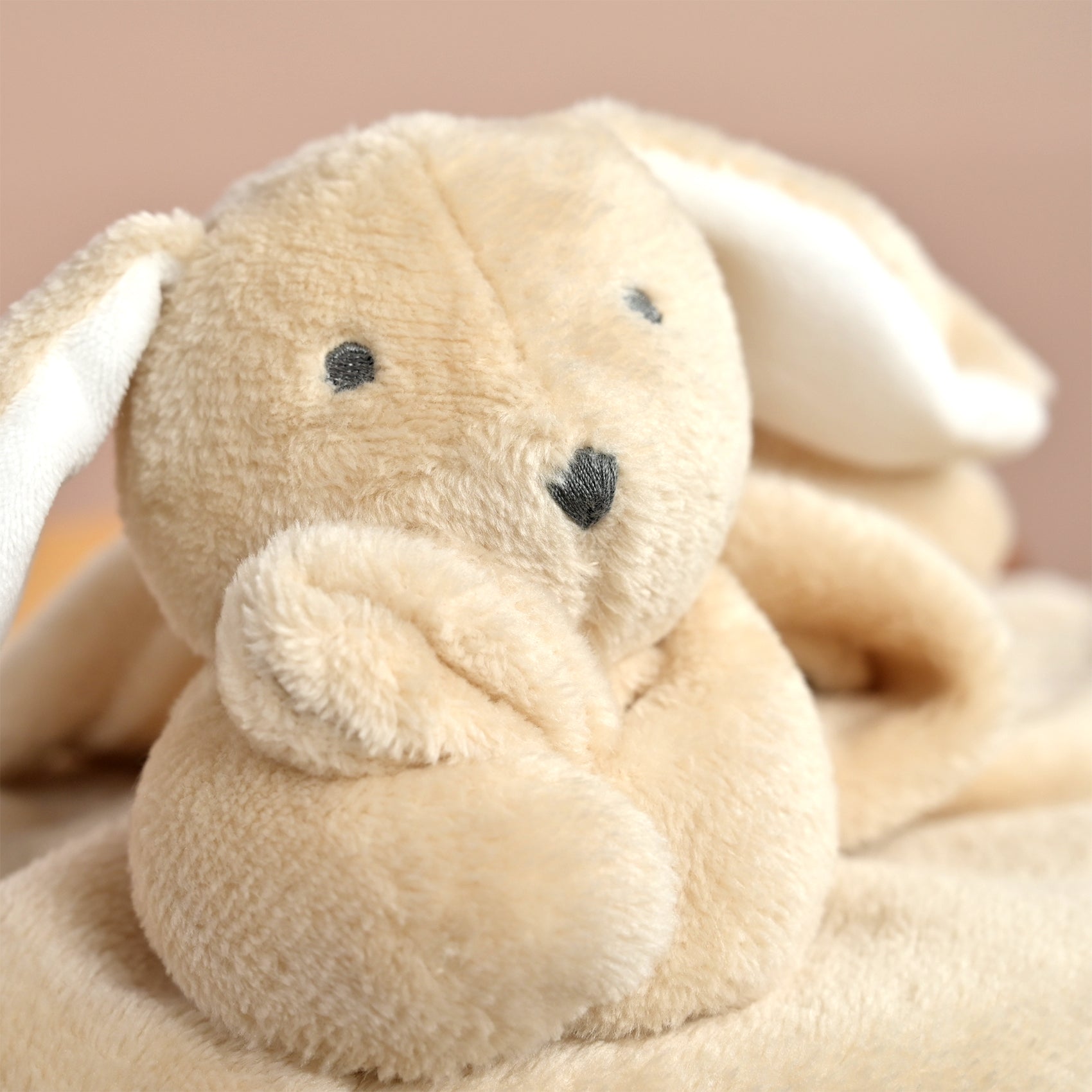 A close-up of a soft plush bunny toy or comforter in warm beige and cream tones, with embroidered black eyes and nose, resting against a pale pink background.