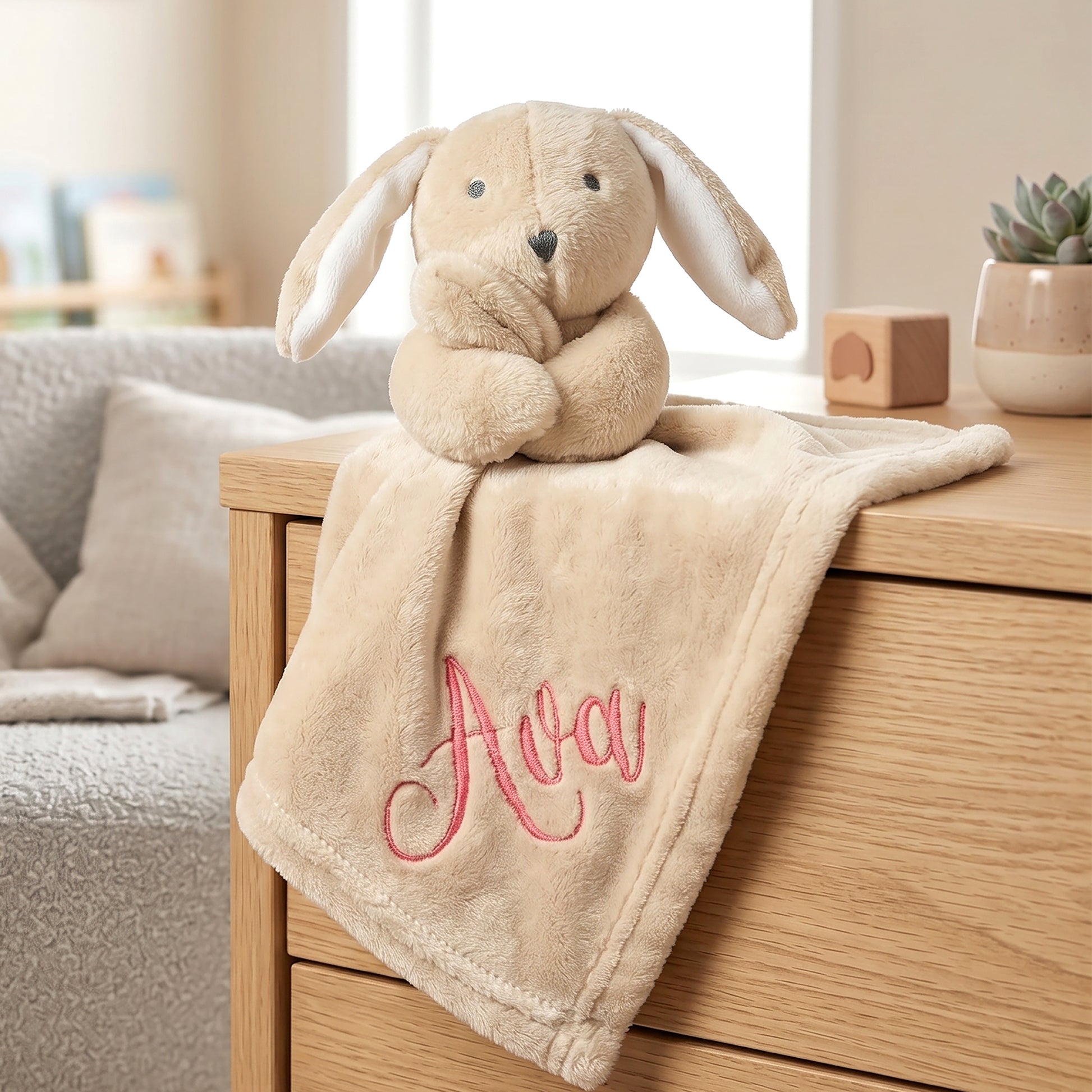 A beige plush bunny comforter with long floppy ears sits on a wooden dresser in a softly lit nursery. A matching beige blanket is draped over the front of the dresser, personalized with pink embroidered script reading 'Ava'.