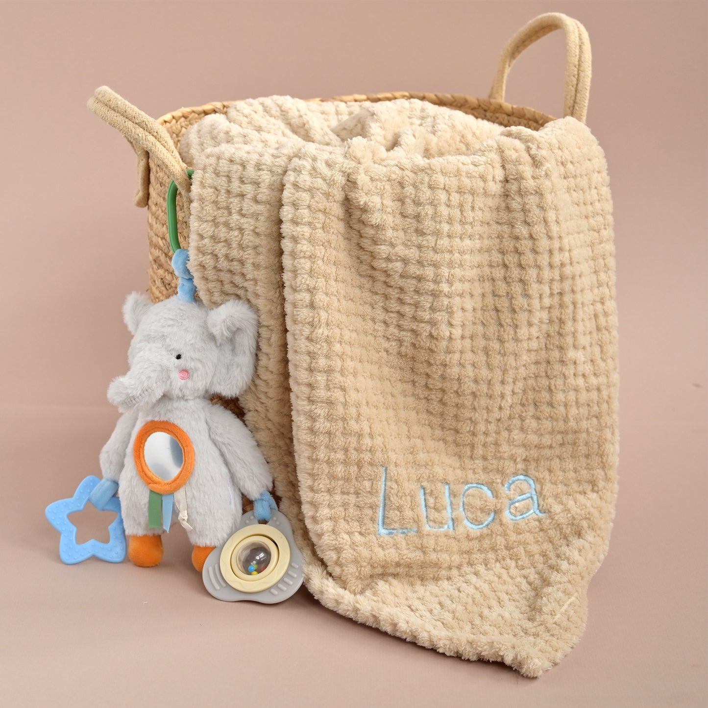A beige textured fabric storage basket with two handles, personalized with the name 'Luca' in light blue embroidery, shown with a plush gray elephant toy and blue star rattle on a solid beige background.