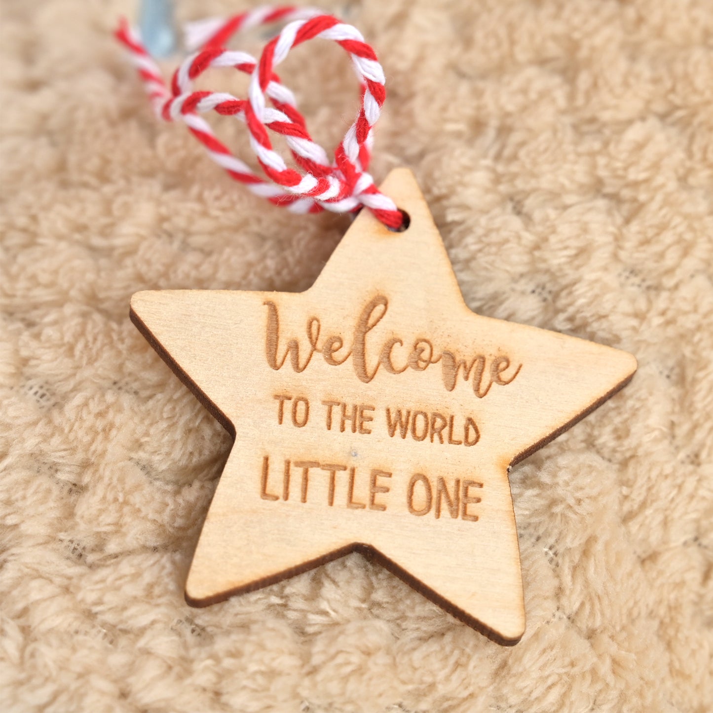 A wooden star-shaped ornament engraved with 'Welcome to the world little one' in a warm natural wood tone, hanging from a red and white twisted string on a soft beige textured blanket.