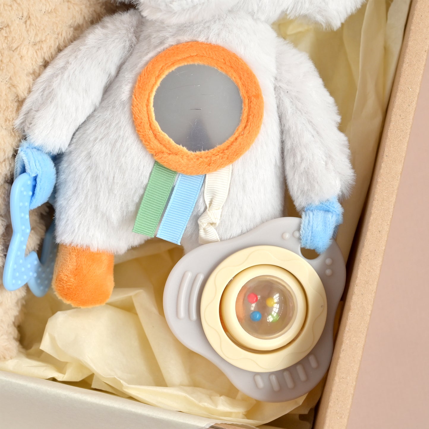 A plush baby gift set inside a box, featuring a white astronaut toy with an orange-trimmed circular mirror, pastel ribbon accents, a blue pacifier clip, and a pacifier with a cream-colored center.
