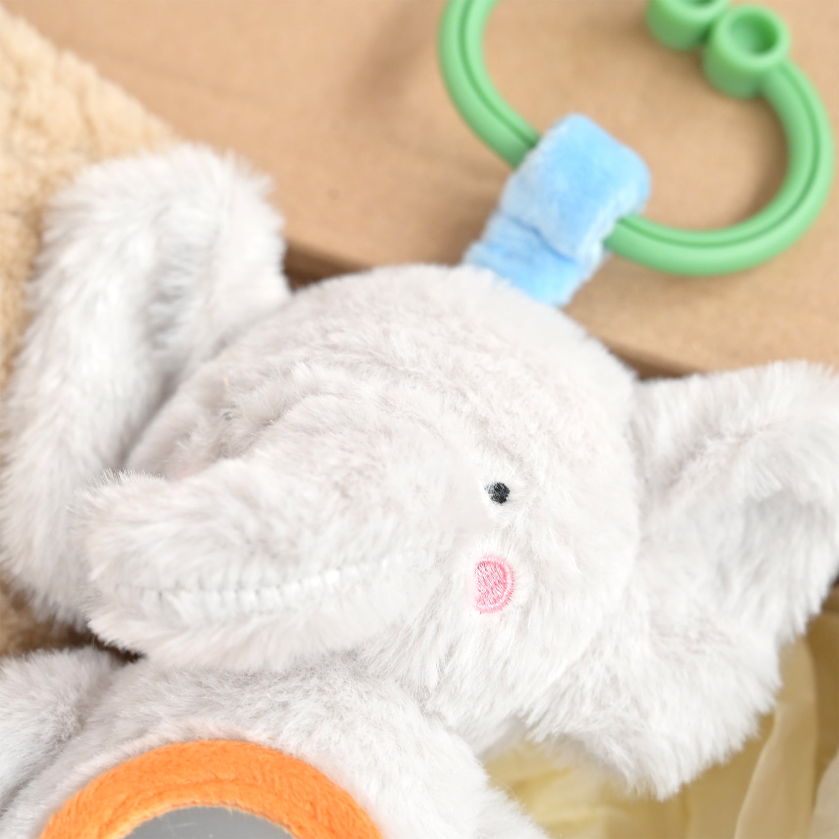 Close-up of a white plush elephant baby toy with a green ring handle, light blue strap, pink embroidered nose, and orange circular accent.