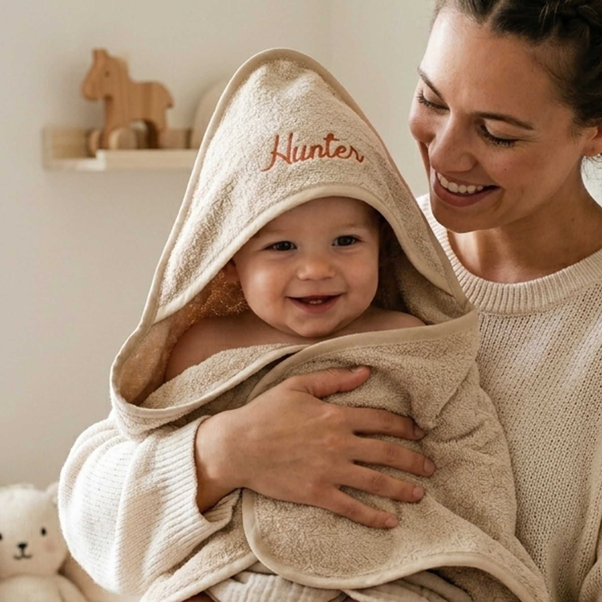 Oatmeal hooded baby towel in cotton terry, personalised with the name Hunter embroidered in terracotta thread, worn by a baby held against a knitted jumper.