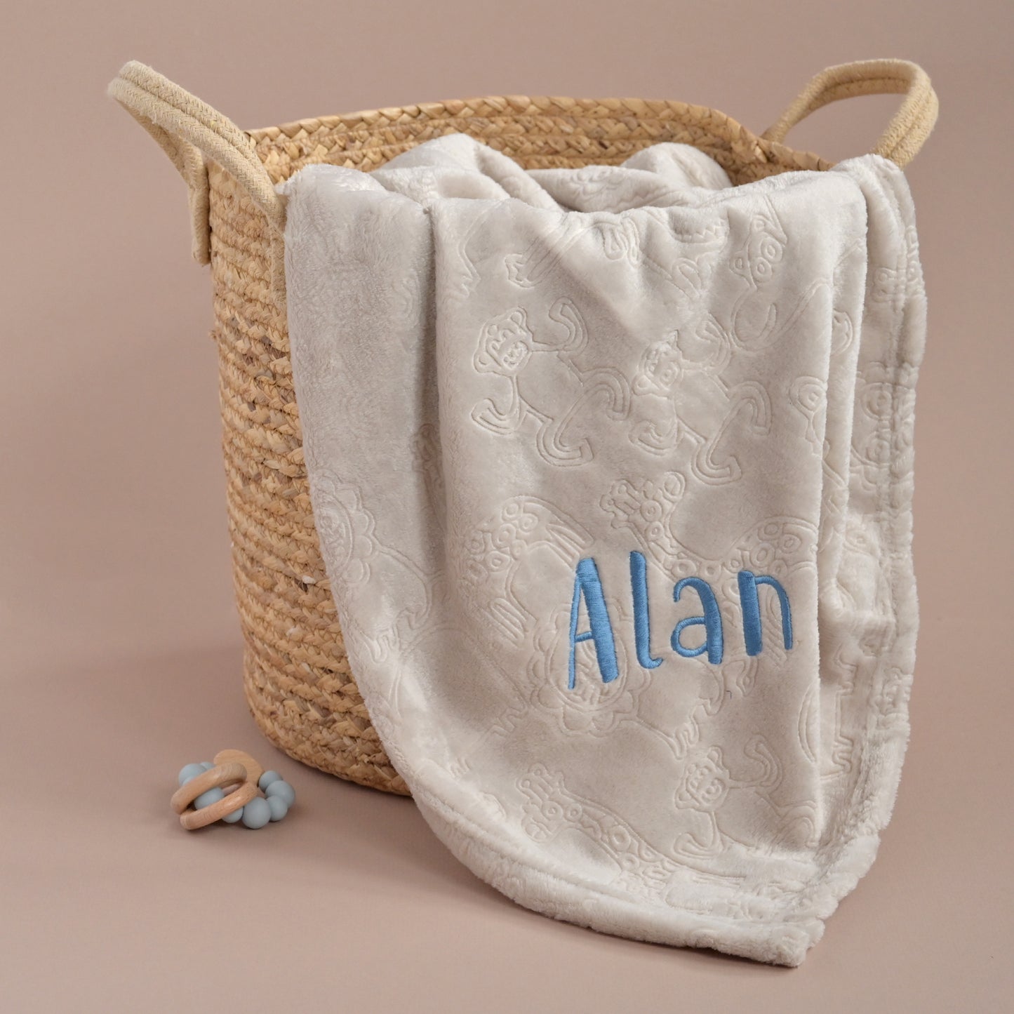 A cream-colored plush baby blanket with a subtle damask pattern and personalized blue embroidery reading 'Alan', draped over a woven straw basket on a solid beige background.