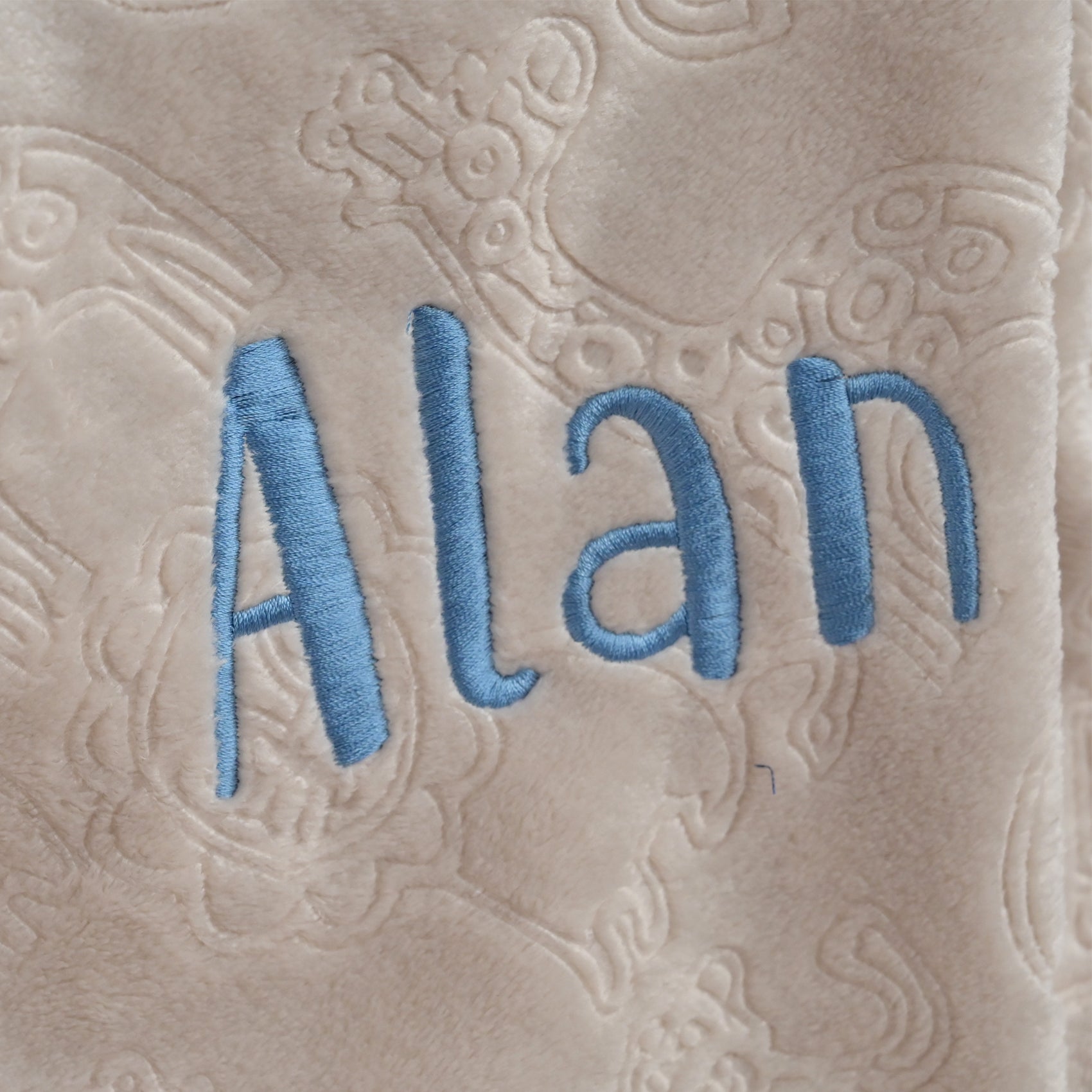 Close-up of a beige plush blanket with an embossed floral pattern and the name 'Alan' embroidered in light blue thread across the center.