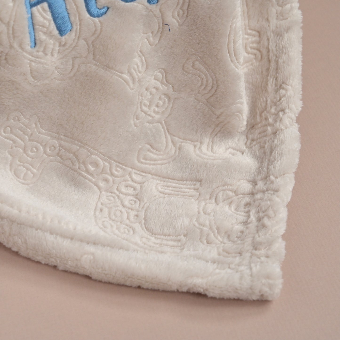 Close-up of a plush cream-colored blanket with a raised embroidered design and light blue lettering, shown on a soft beige background.